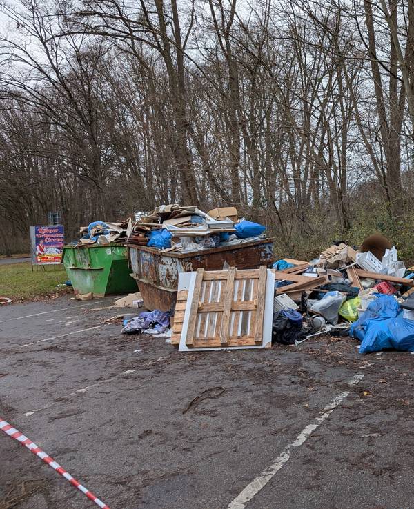 Auf einem Parkplatz stehen zwei übervolle Müllcontainer, daneben liegt weiterer Müll auf dem Boden.