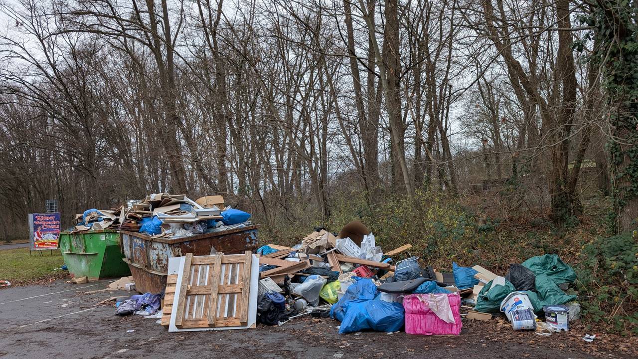 Auf einem Parkplatz stehen zwei übervolle Müllcontainer, daneben liegt weiterer Müll auf dem Boden.