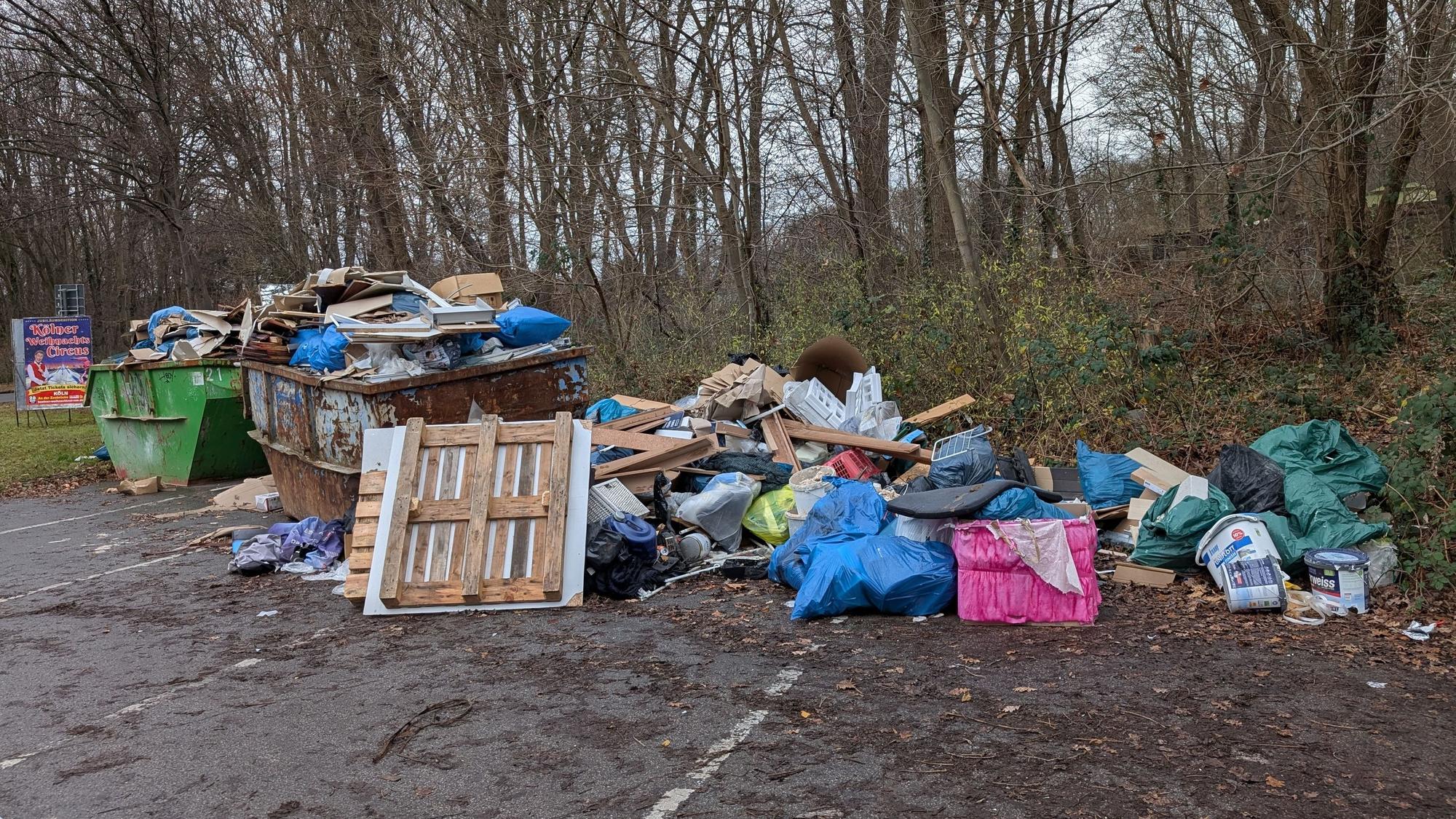 Auf einem Parkplatz stehen zwei übervolle Müllcontainer, daneben liegt weiterer Müll auf dem Boden.