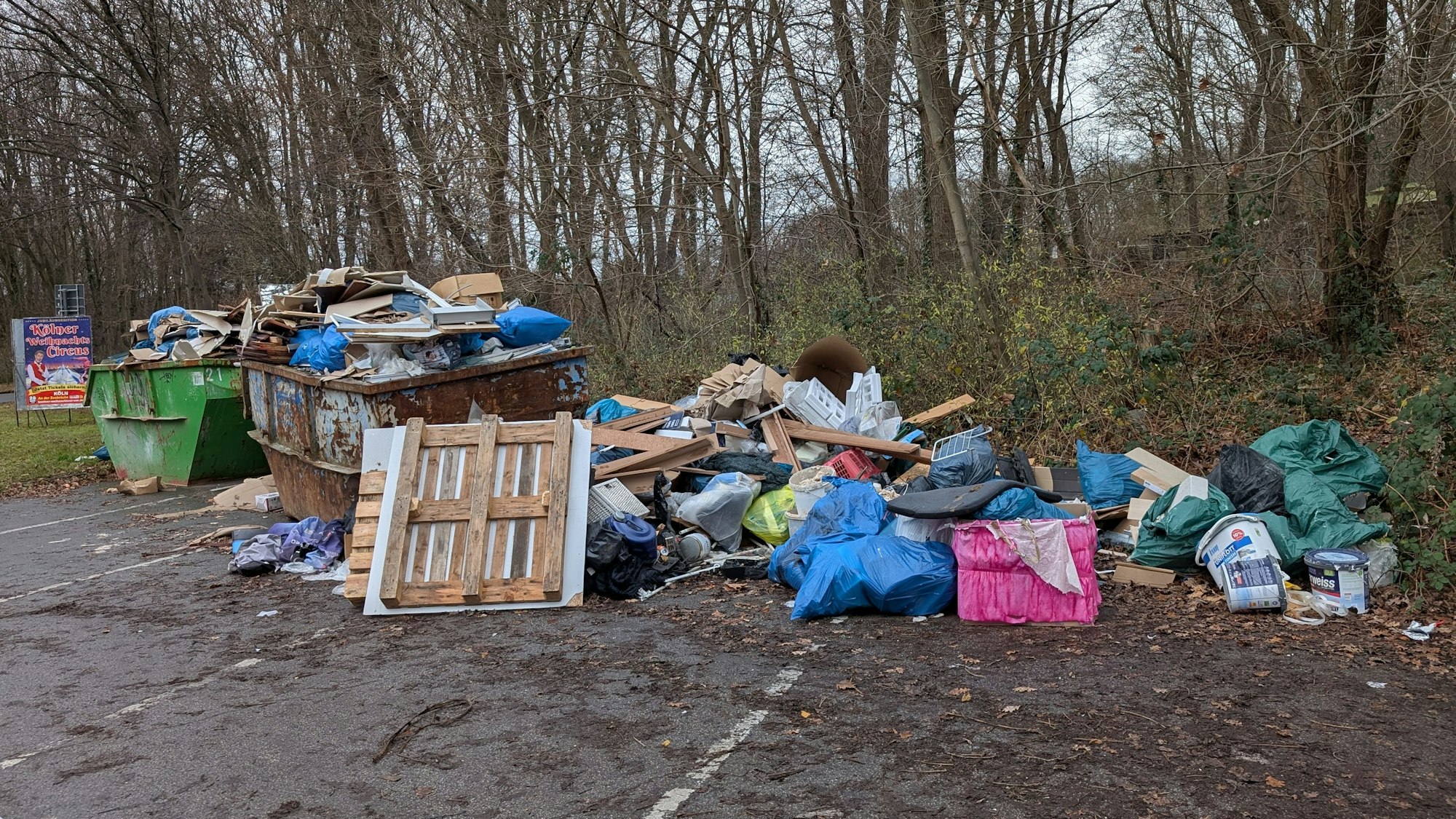 Auf einem Parkplatz stehen zwei übervolle Müllcontainer, daneben liegt weiterer Müll auf dem Boden.