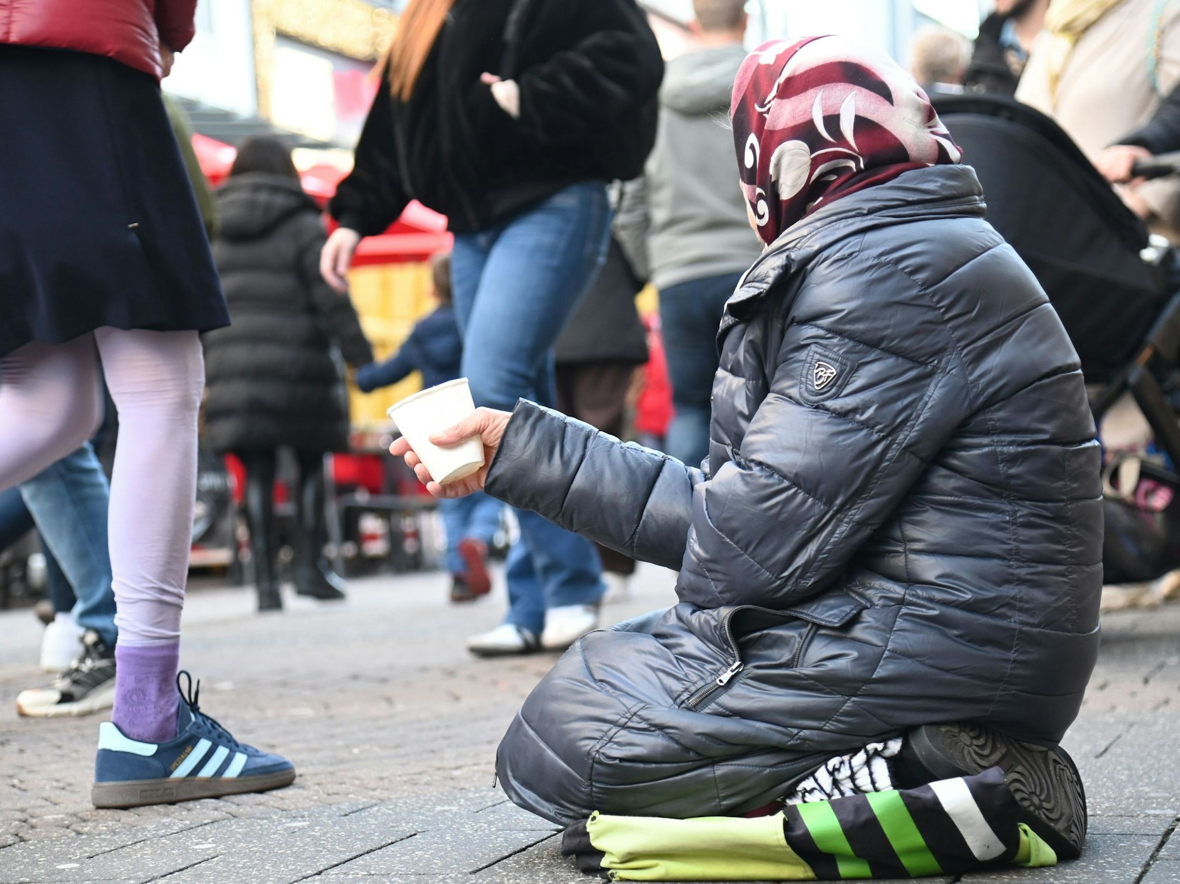 Eine Bettlerin kniet auf einer Unterlage in einer Einkaufsstraße und hält einen Becher vor sich, während Menschen an ihr vorbeilaufen.