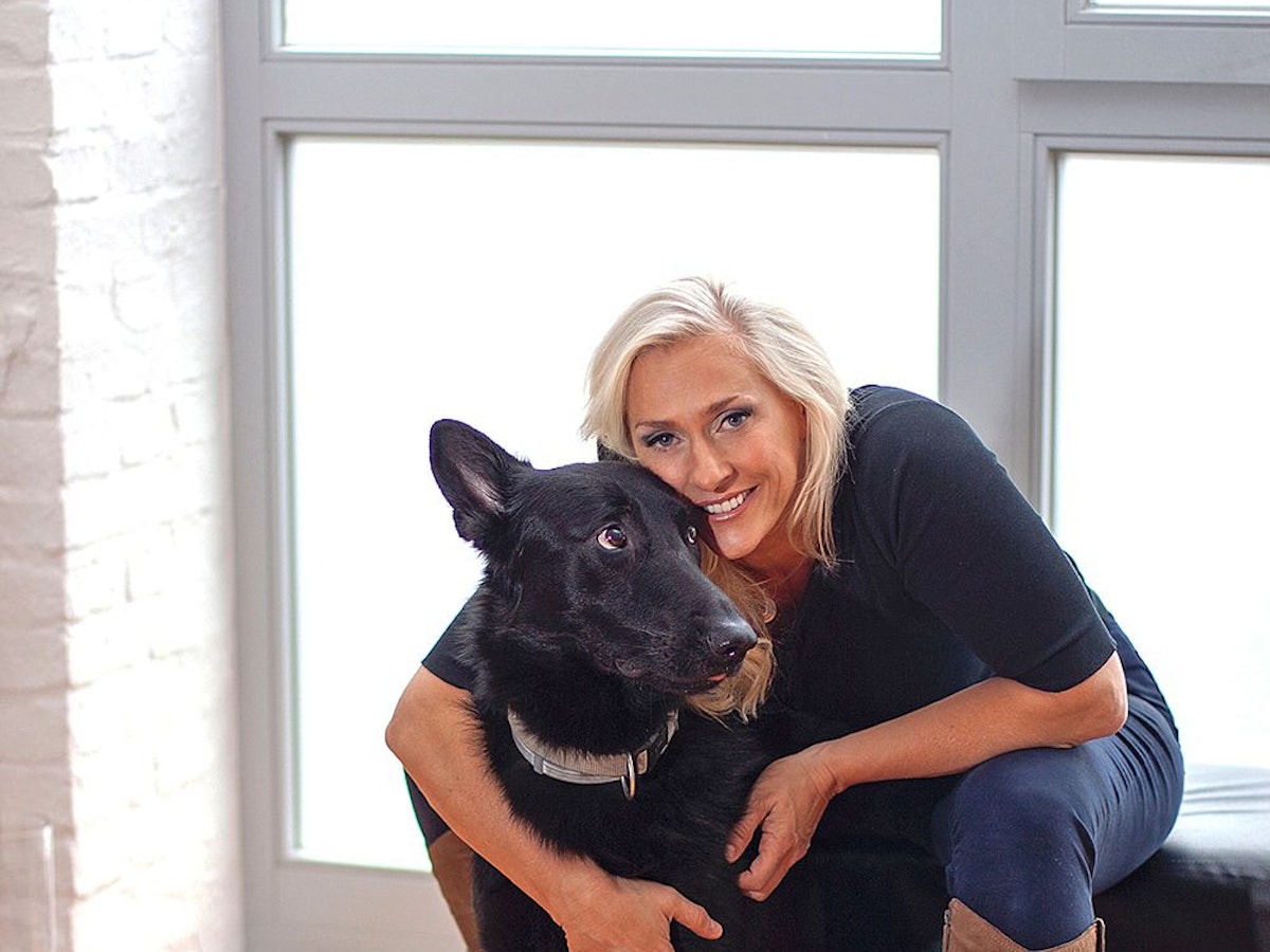 Eine blonde Frau sitzt vor einem Fenster und hat den Arm um einen schwarzen Hund gelegt.