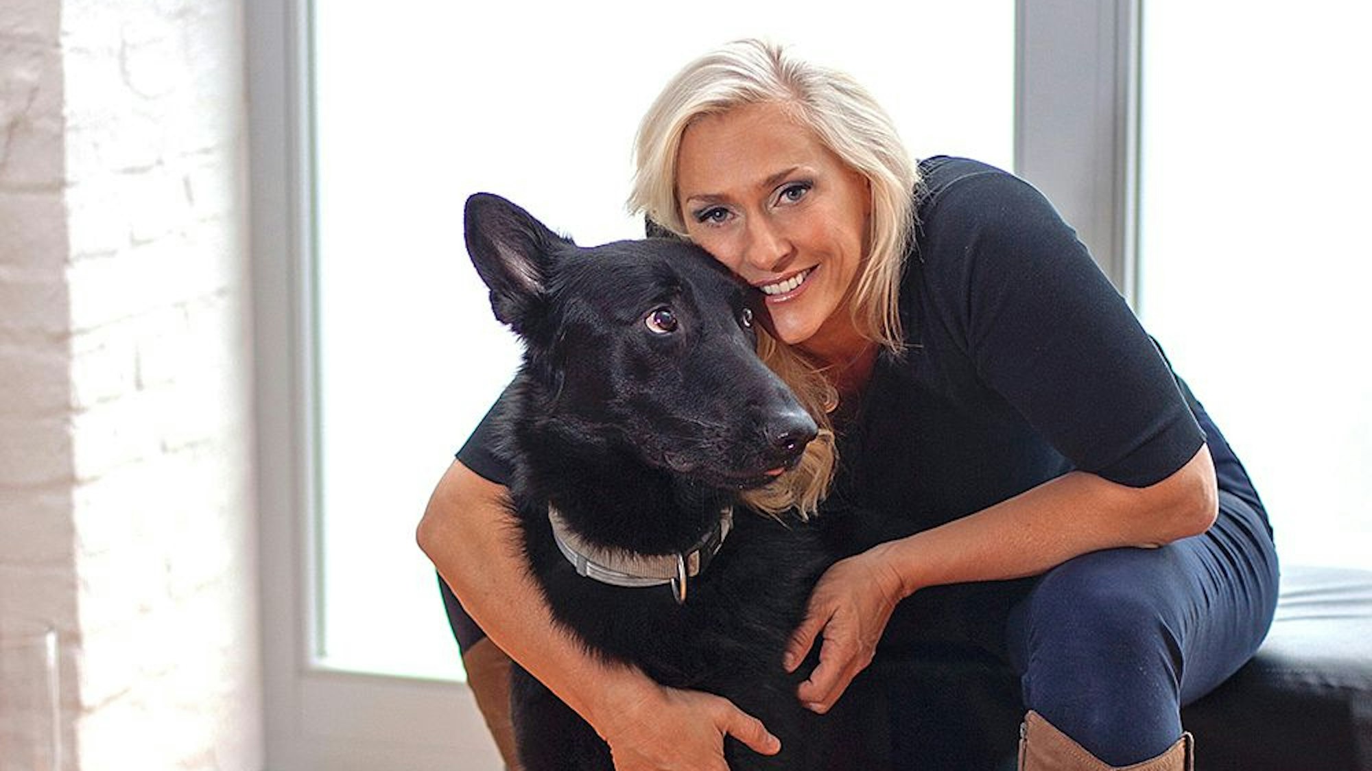Eine blonde Frau sitzt vor einem Fenster und hat den Arm um einen schwarzen Hund gelegt.