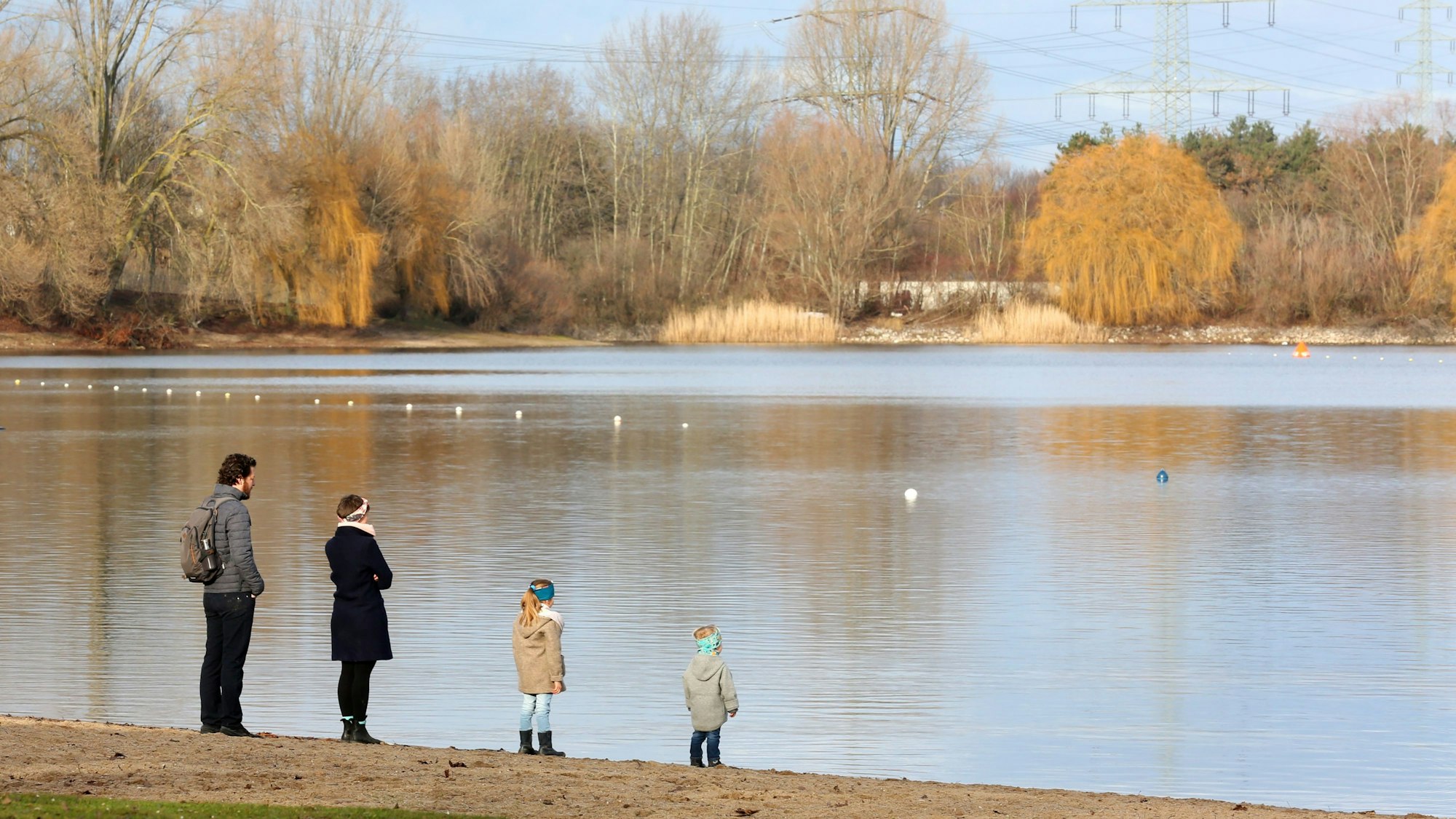Zwei Erwachsene und zwei Kinder stehen an einem Wintertag am Ufer eines Sees und blicken übers Wasser.