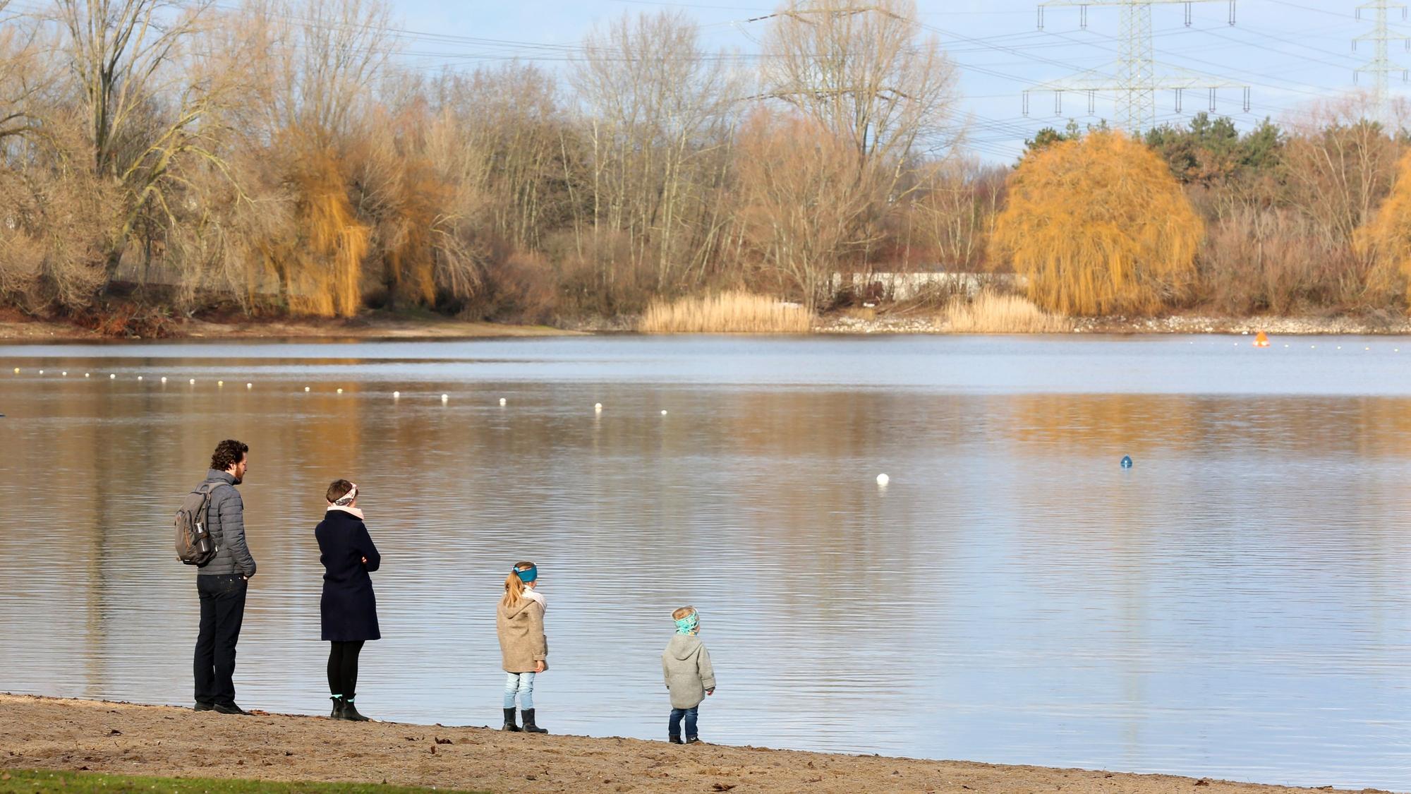 Zwei Erwachsene und zwei Kinder stehen an einem Wintertag am Ufer eines Sees und blicken übers Wasser.