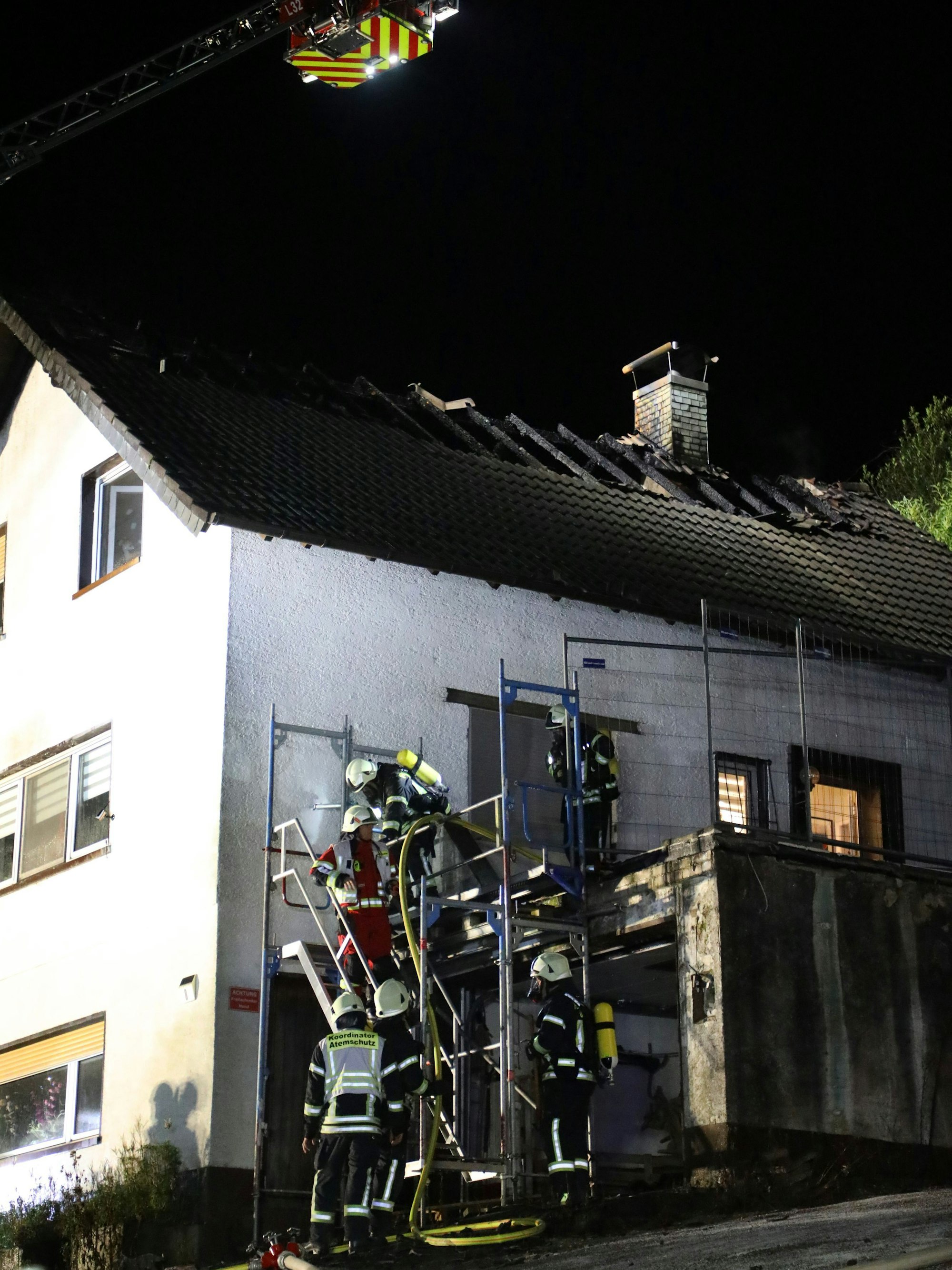 Feuerwehrleute steigen über ein Gerüst zu einem Haus hinauf. Das Dach des Hauses ist abgebrannt.