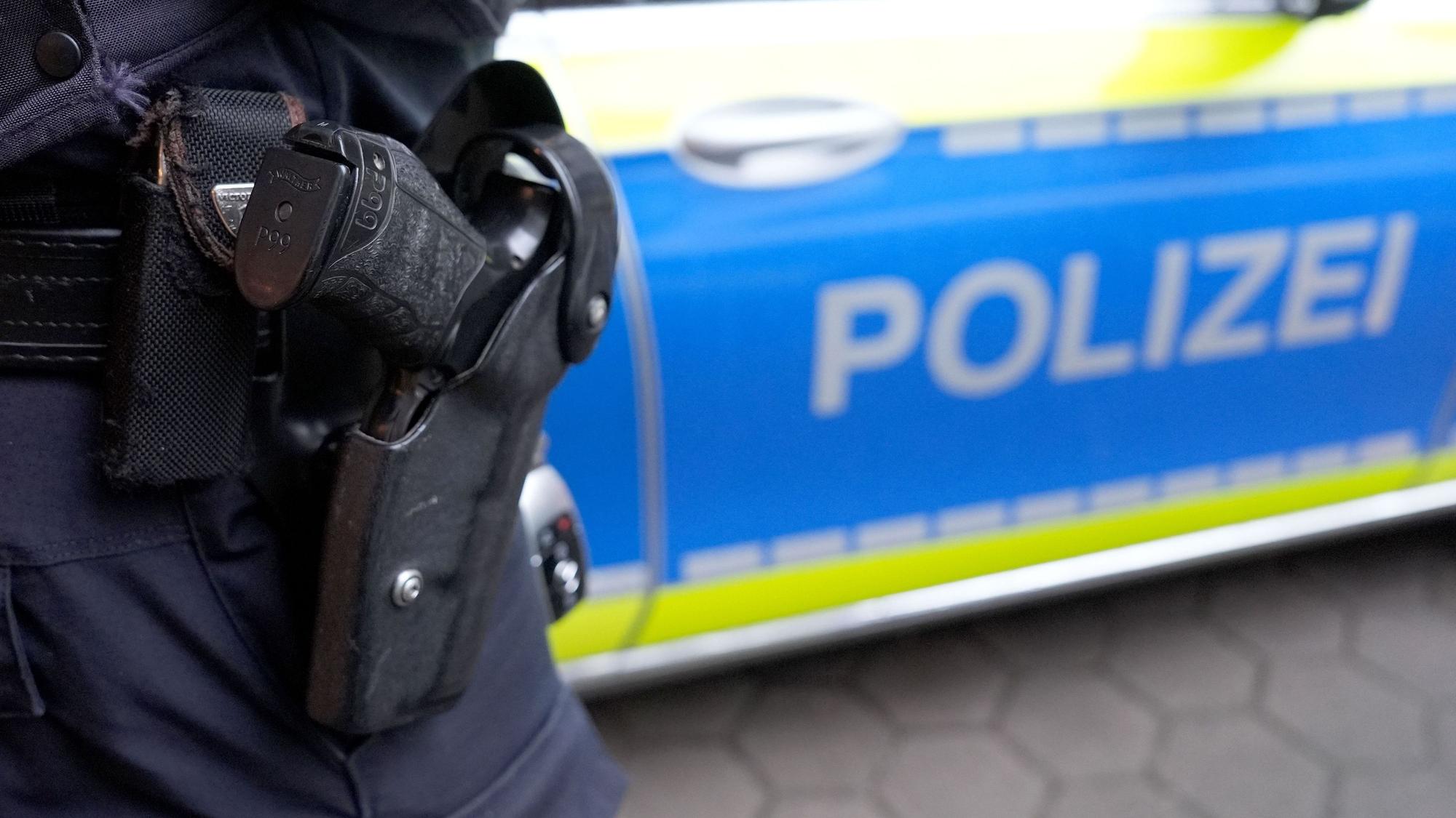 Das Symbolfoto zeigt einen Polizisten mit Schusswaffe vor einem Einsatzfahrzeug der Polizei.