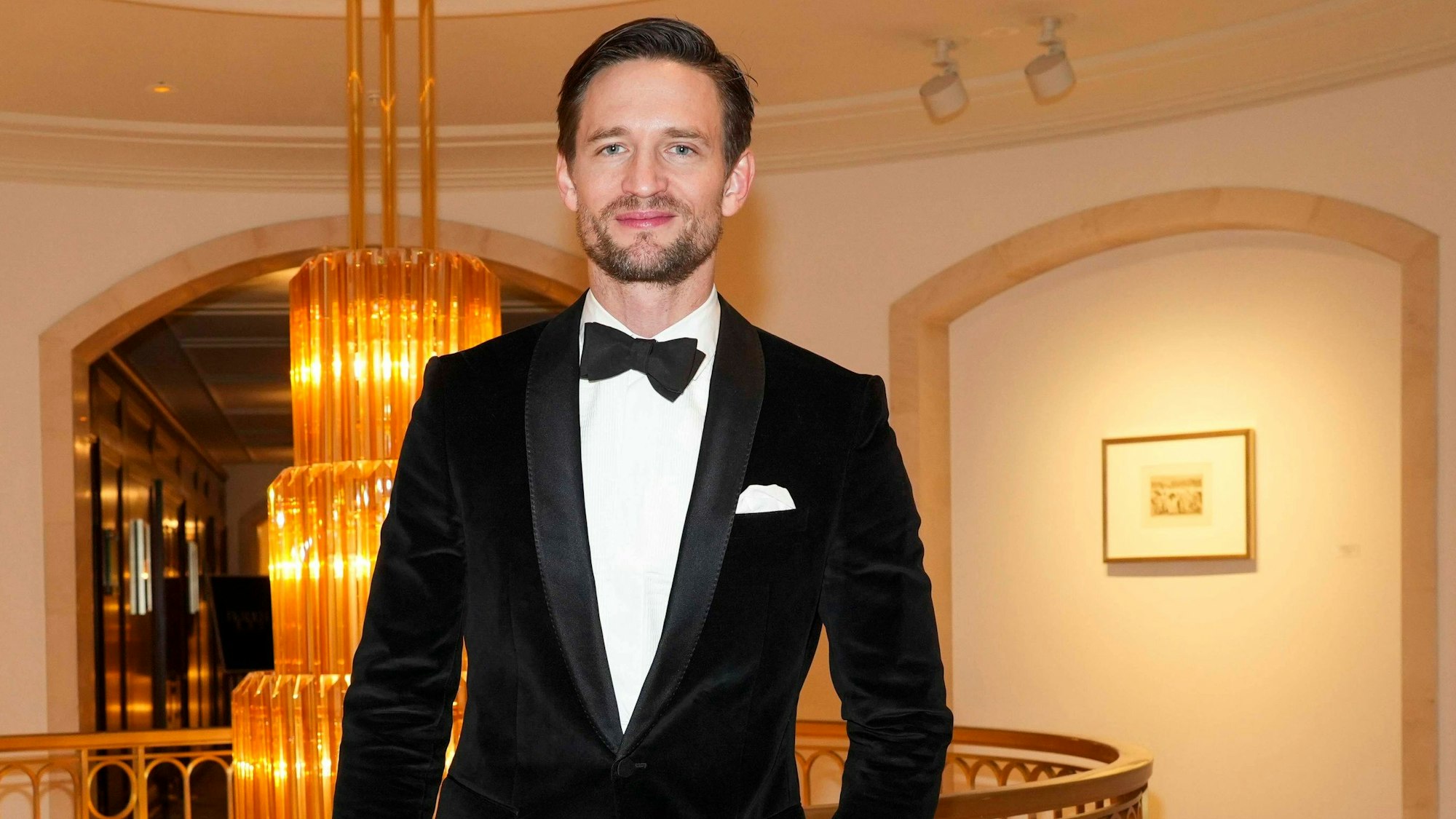 Ein junger Mann mit gegelten Haaren und akkurat sitzendem schwarzen Anzug mit Einstecktuch und Fliege schaut in die Kamera