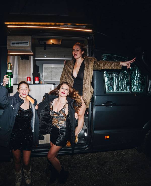 Drei festlich gekleidete Frauen stehen vor einem Campingbus und prosten sich mit Champagner zu.