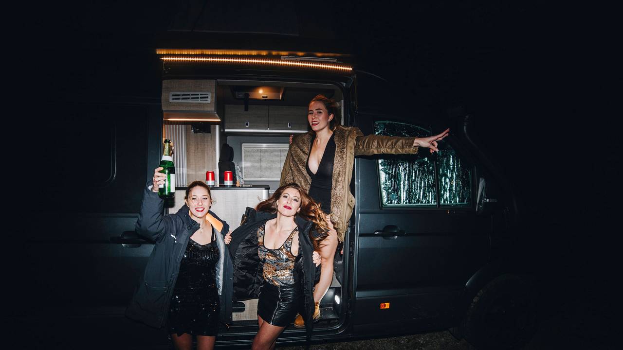 Drei festlich gekleidete Frauen stehen vor einem Campingbus und prosten sich mit Champagner zu.