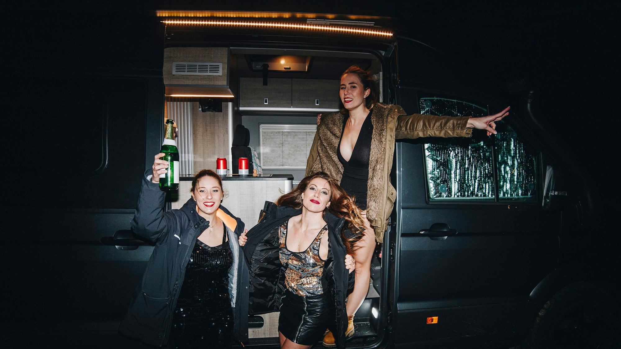Drei festlich gekleidete Frauen stehen vor einem Campingbus und prosten sich mit Champagner zu.