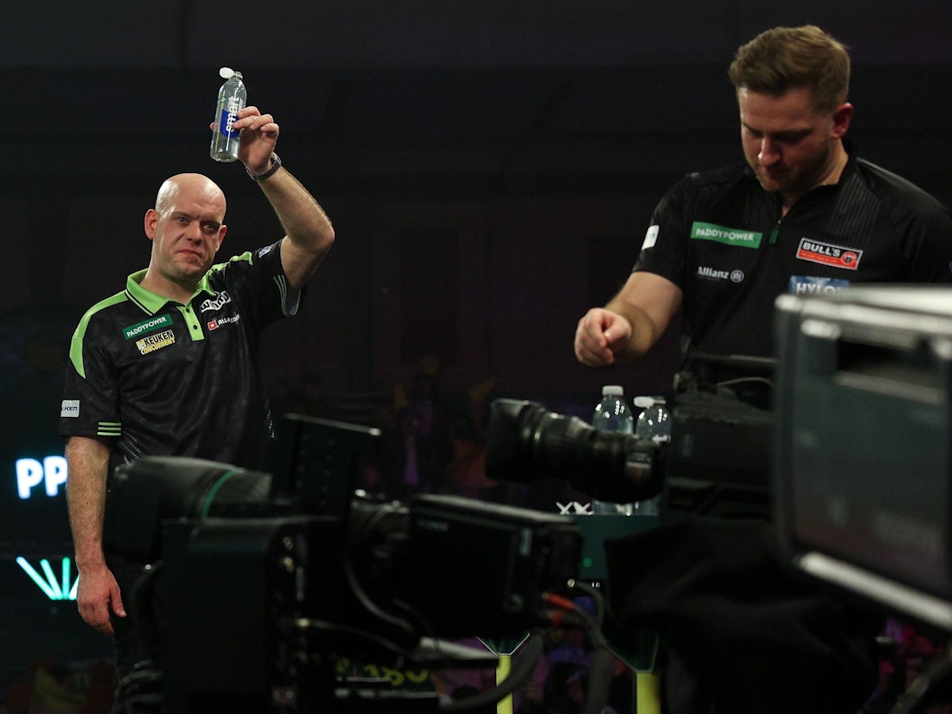 Michael van Gerwen hält im Hintergrund eine Wasserflasche in die Luft während sich im Vordergrund Arno Merk auf das Match vorbereitet.