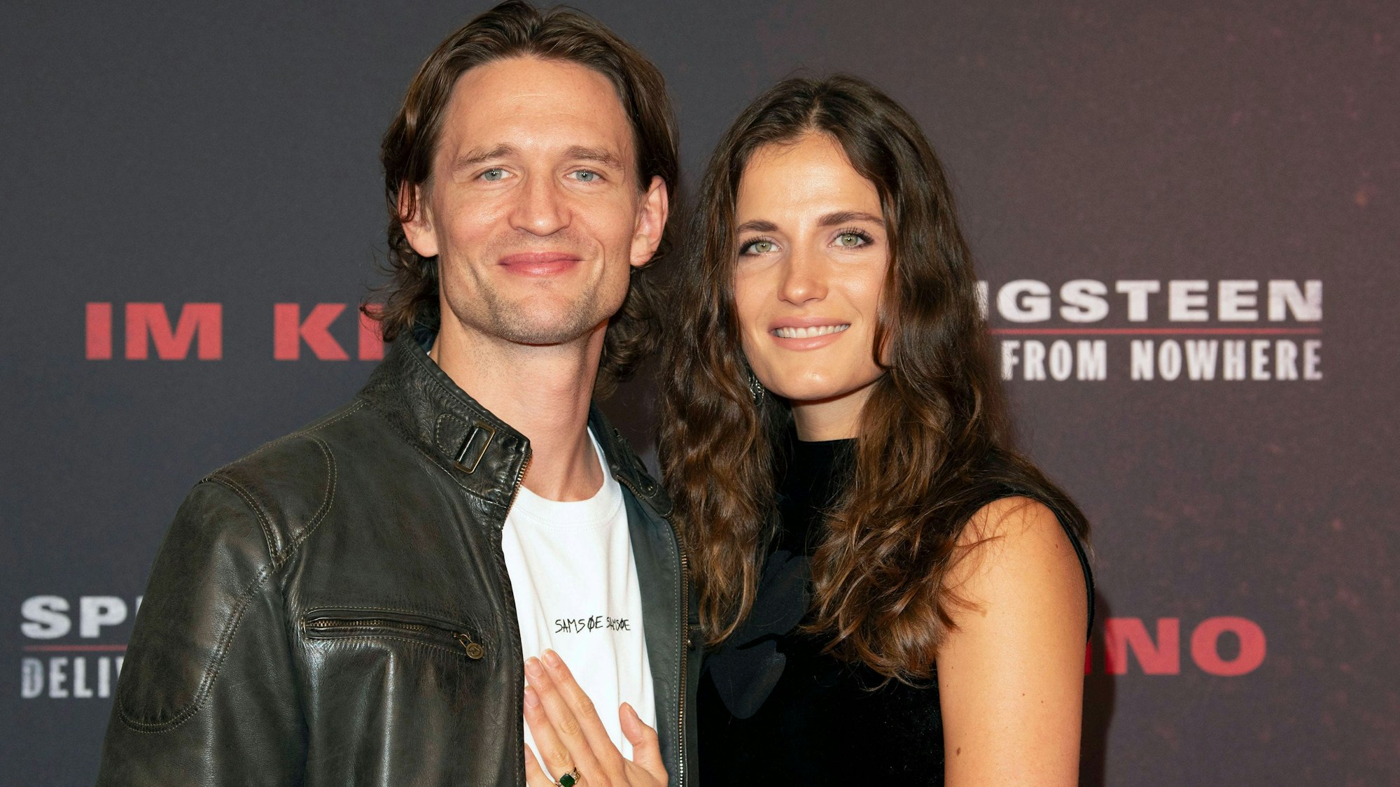 Ein dunkelhaariger Mann und eine brünette Frau mit langen Haaren halten sich um Arm und schauen freundlich drein