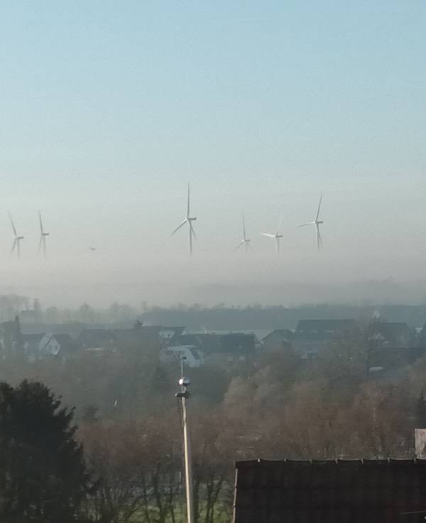 Blick über eine Stadt auf Windräder, von denen wegen Nebels nur die oberen Rotorblätter zu sehen sind und sie daher zu schweben scheinen.
