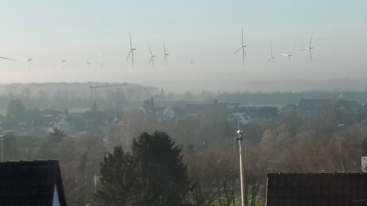 Blick über eine Stadt auf Windräder, von denen wegen Nebels nur die oberen Rotorblätter zu sehen sind und sie daher zu schweben scheinen.