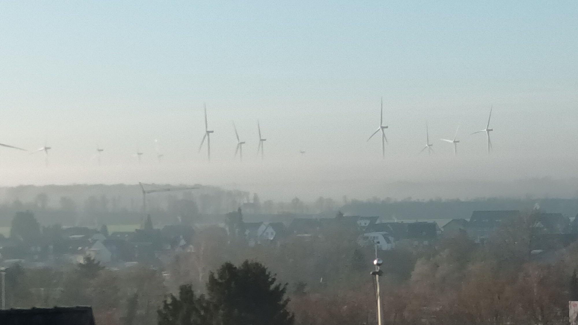Blick über eine Stadt auf Windräder, von denen wegen Nebels nur die oberen Rotorblätter zu sehen sind und sie daher zu schweben scheinen.