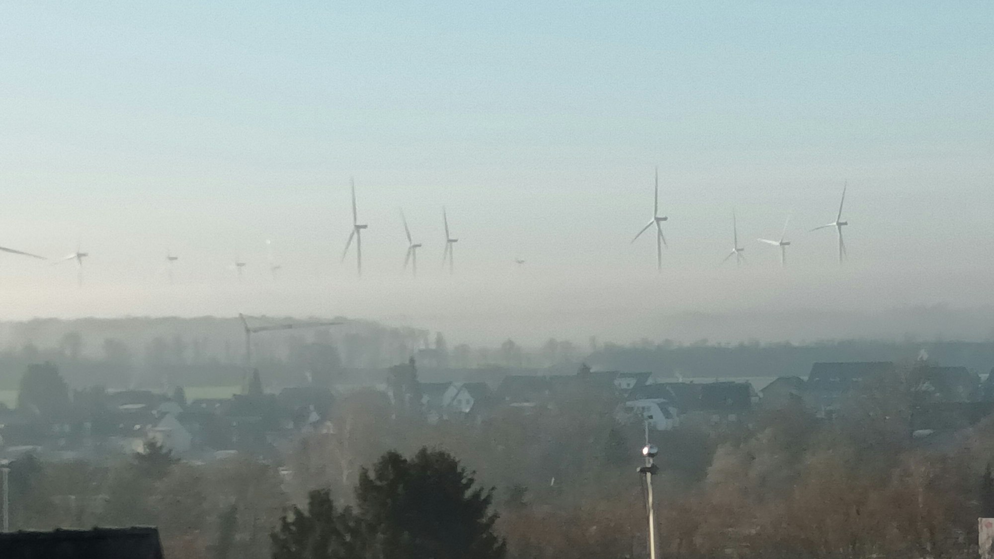 Blick über eine Stadt auf Windräder, von denen wegen Nebels nur die oberen Rotorblätter zu sehen sind und sie daher zu schweben scheinen.
