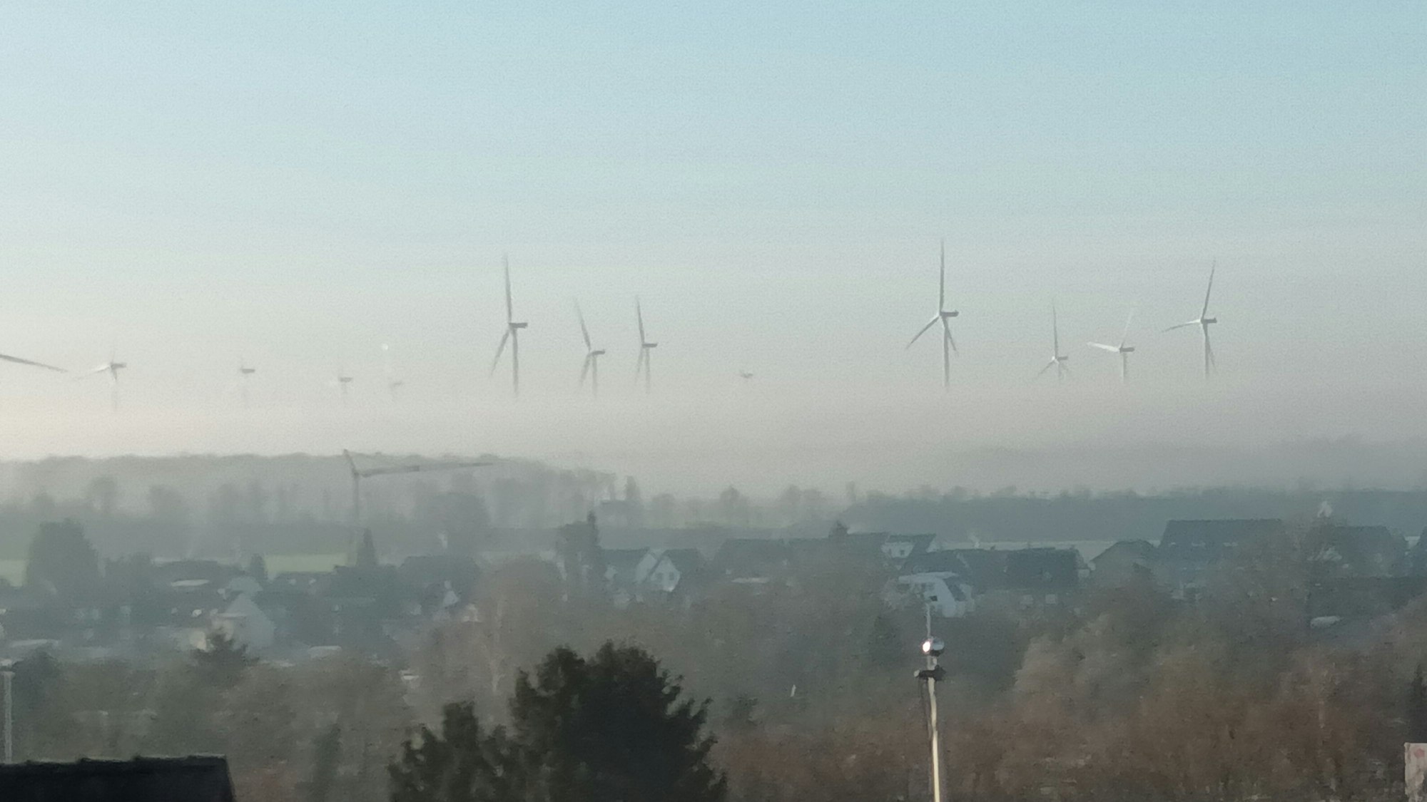 Blick über eine Stadt auf Windräder, von denen wegen Nebels nur die oberen Rotorblätter zu sehen sind und sie daher zu schweben scheinen.