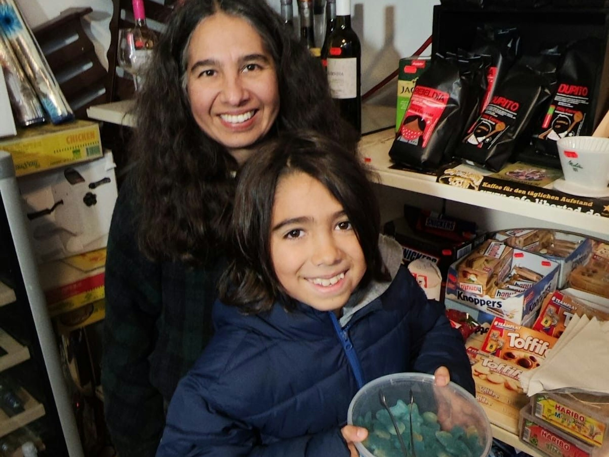 Eine Mutter und ihr Sohn sind in einem Kiosk, der Junge hält eine Schale mit Süßigkeiten vor sich.