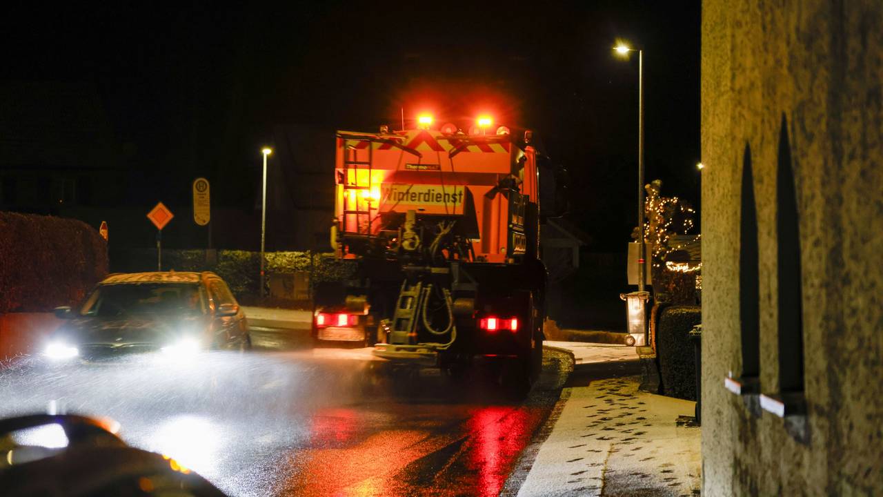 Ein Streu- und Räumfahrzeug ist im Dunkeln auf einer Straße unterwegs. Auf dem Gehweg liegt eine dünne Schneeschicht.