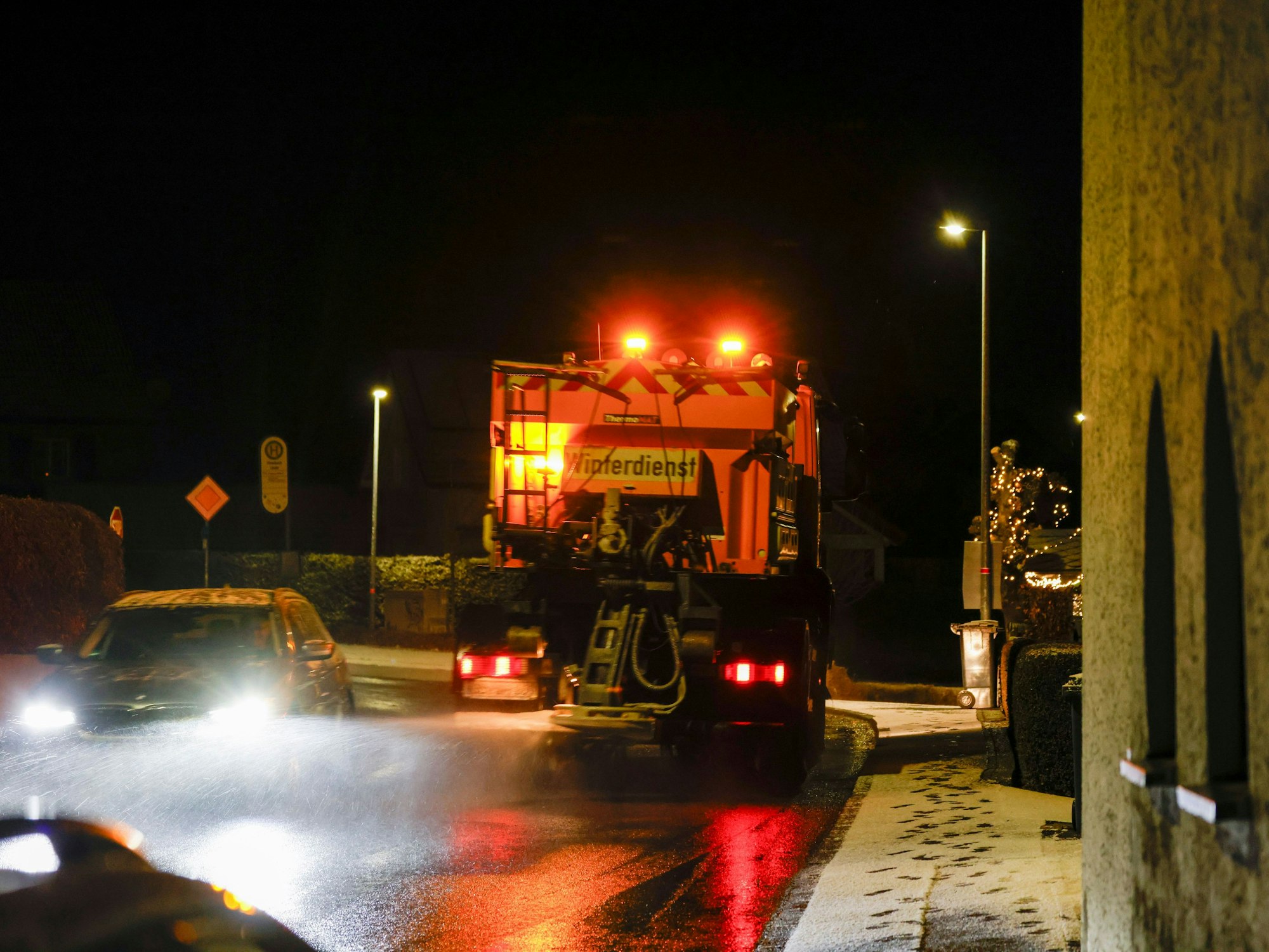 Ein Streu- und Räumfahrzeug ist im Dunkeln auf einer Straße unterwegs. Auf dem Gehweg liegt eine dünne Schneeschicht.