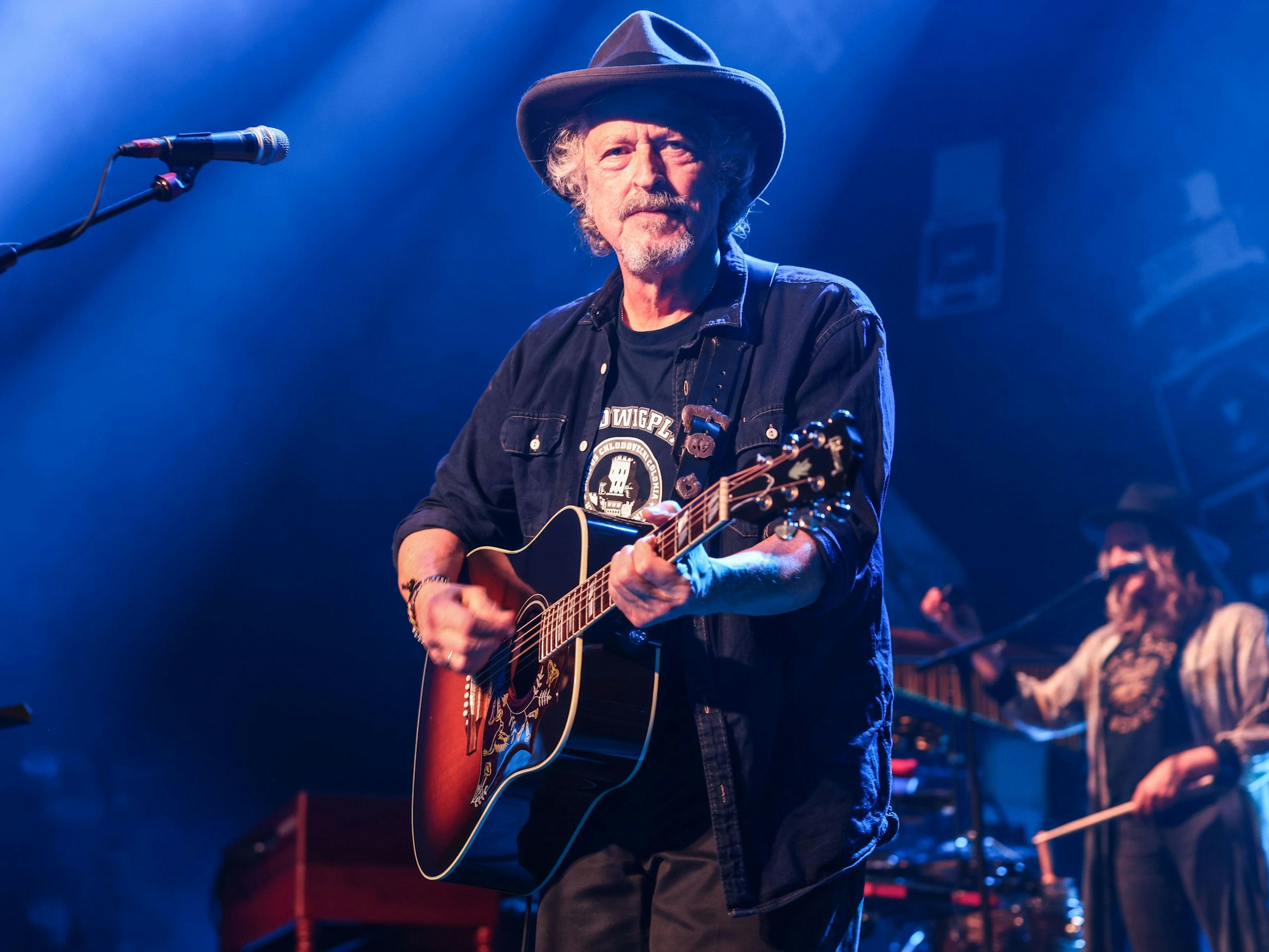 Wolfgang Niedecken steht mit Gitarre und Hut auf einer Bühne.
