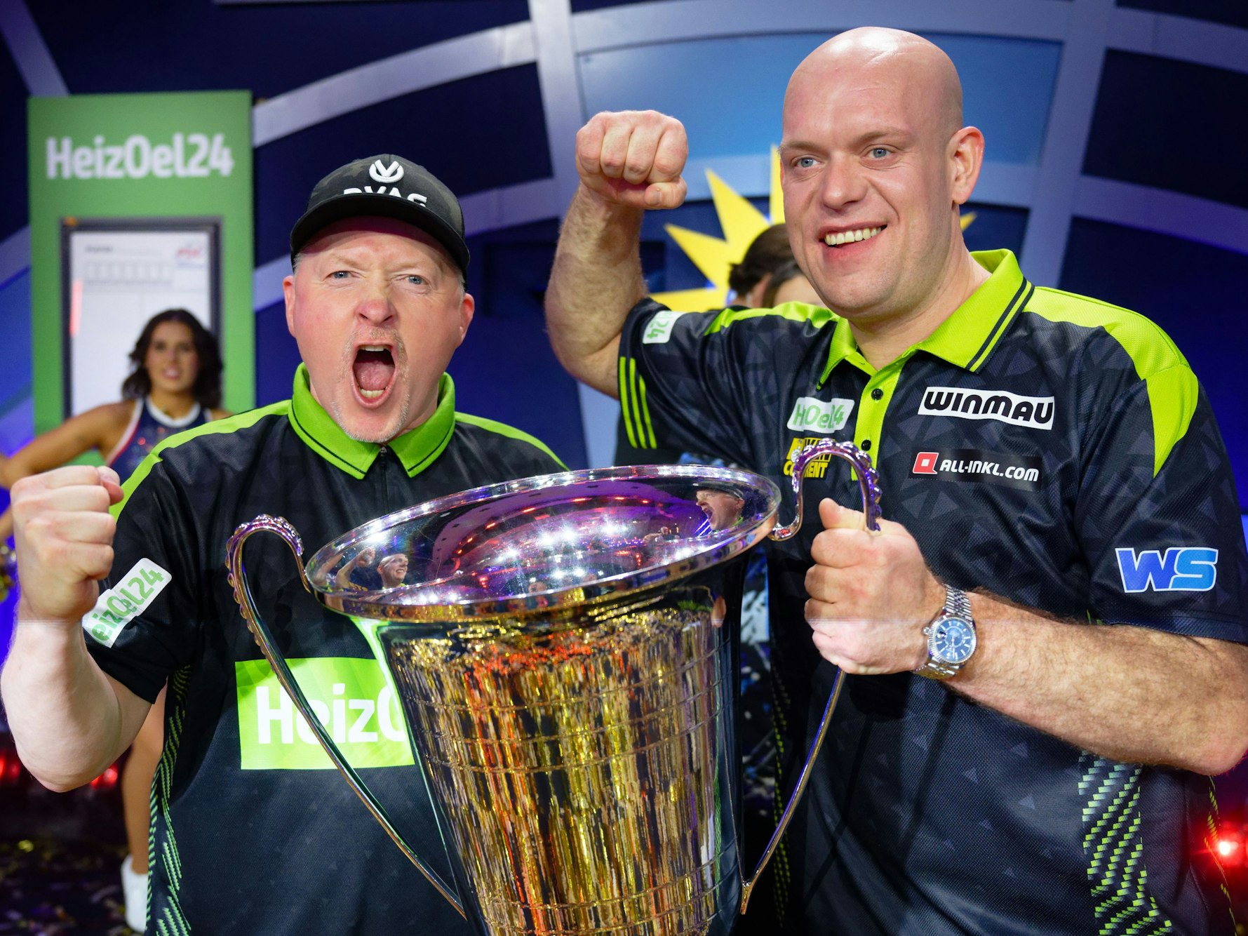 Joey Kelly und Michael van Gerwen stehen bei der Promi-Darts-WM 2026 mit dem Siegerpokal auf der Bühne.