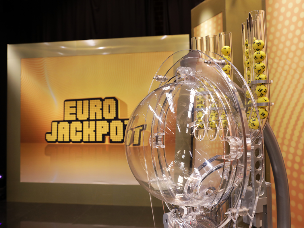 Das Bild zeigt die moderne Ziehungsmaschine für die Eurojackpot-Lotterie mit gelben Kugeln im Vordergrund. Im Hintergrund ist groß das goldene „EURO JACKPOT“-Logo auf einer beleuchteten Wand zu sehen.