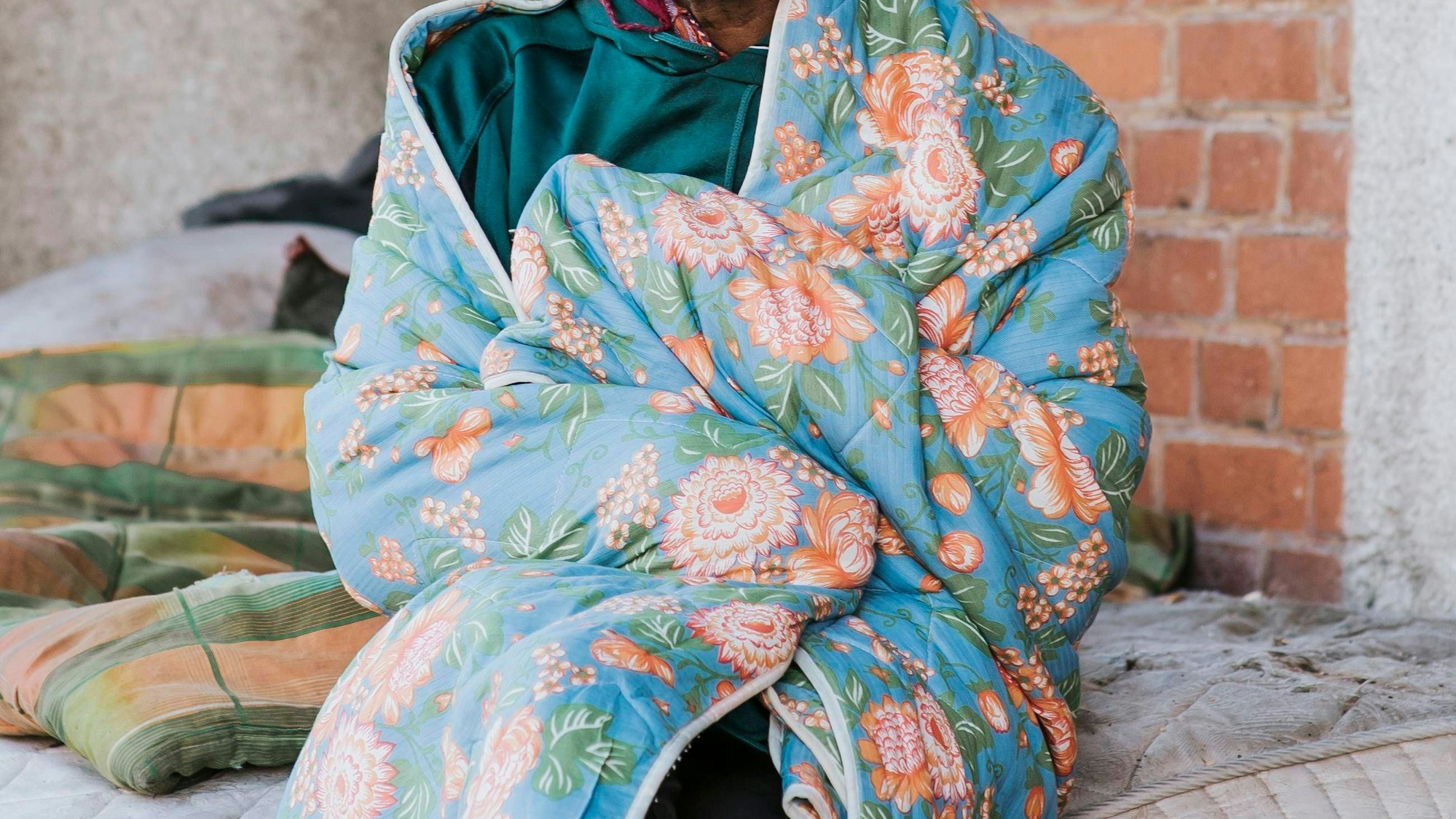 Obdachloser Mann sitzt auf Matratze mit Decke