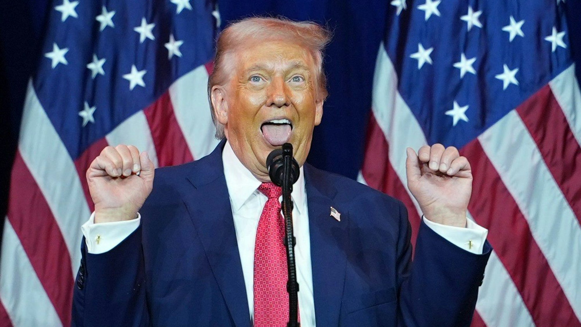 Präsident Donald Trump hält die Hände hoch, als würde er etwas stemmen, dazu streckt er die Zunge raus.