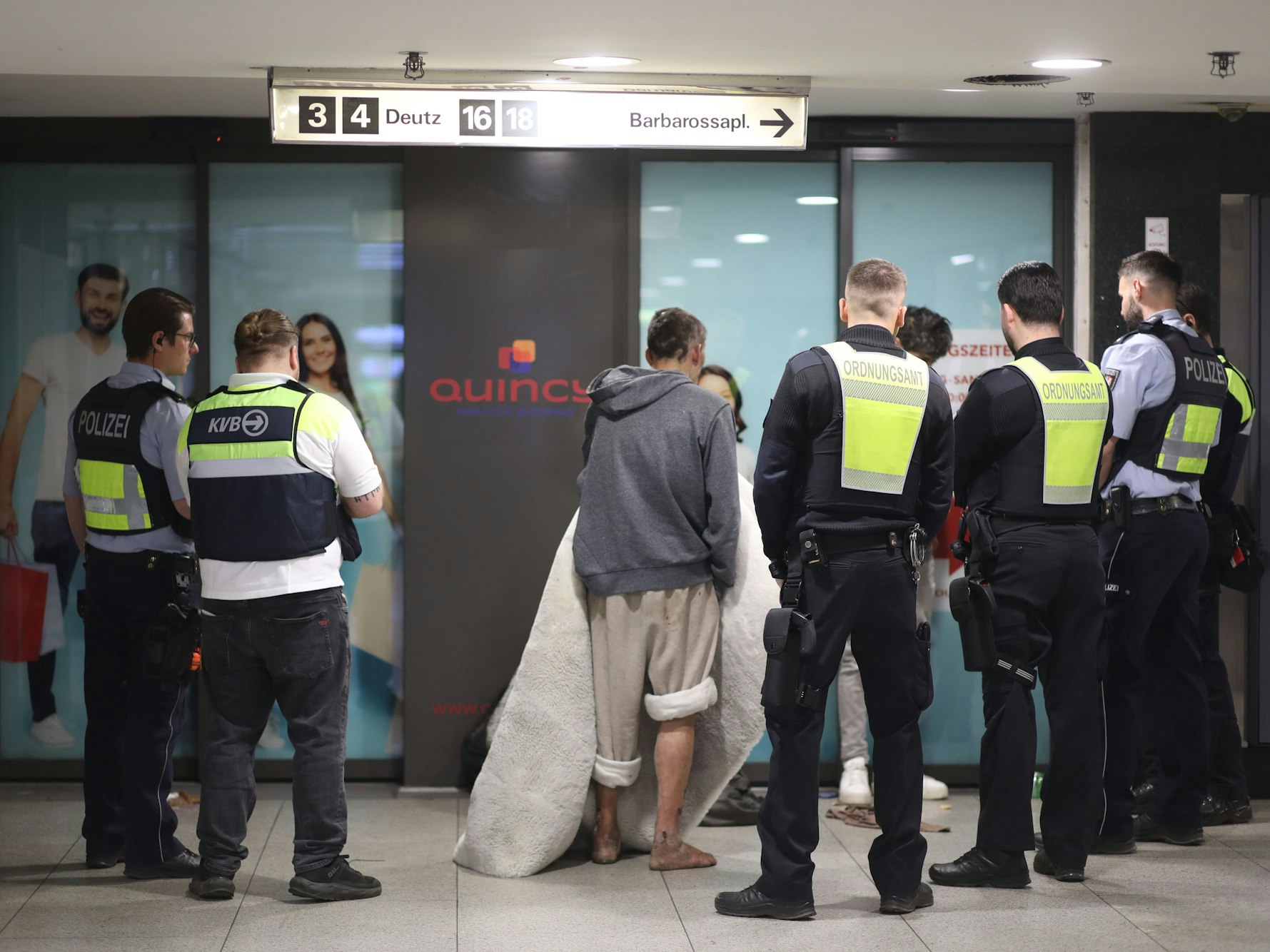 Kräfte von Polizei, Stadt und KVB stehen um Personen herum, darunter ein Mann, der barfuß ist.