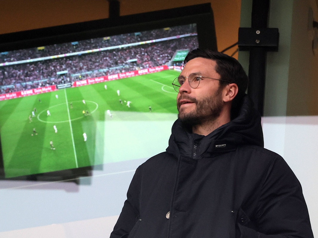 Jonas Hector steht vor einem Fernseher, auf dem ein Fußballspiel läuft.