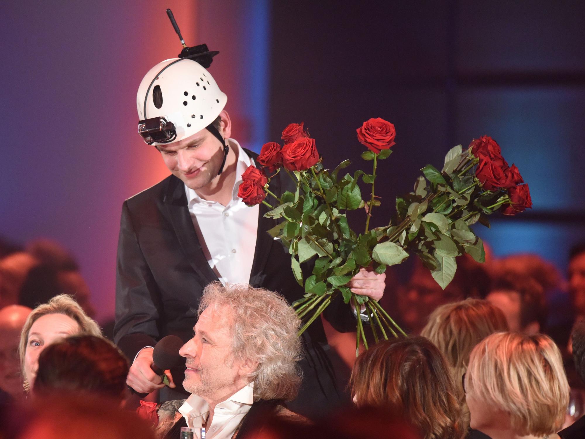 Ein Mann mit Helm und Kamera überreicht einem Mann im Publikum einen Rosenstrauß