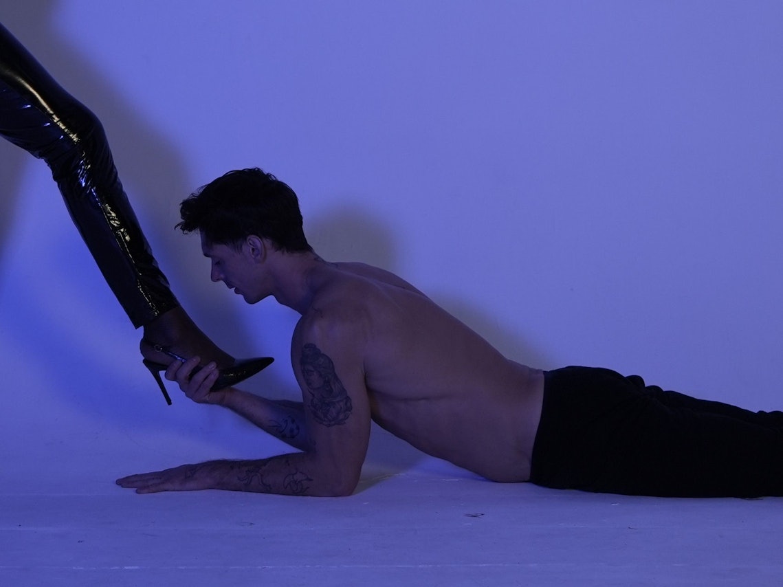 Ein junger Mann liegt auf dem Boden und hält den Fuß mit High Heels einer Dame mit Lack-Lederhose in der Hand.