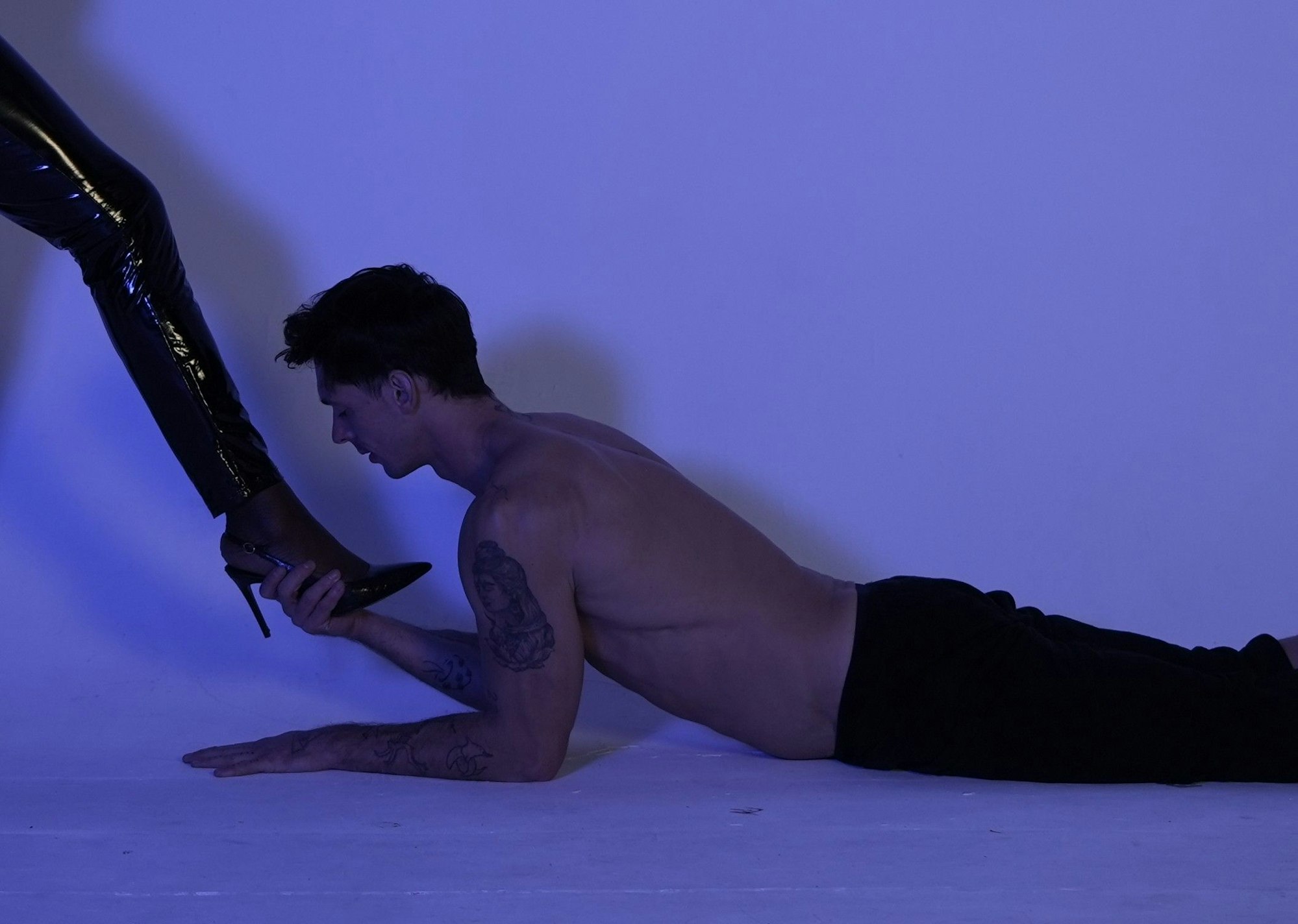 Ein junger Mann liegt auf dem Boden und hält den Fuß mit High Heels einer Dame mit Lack-Lederhose in der Hand.