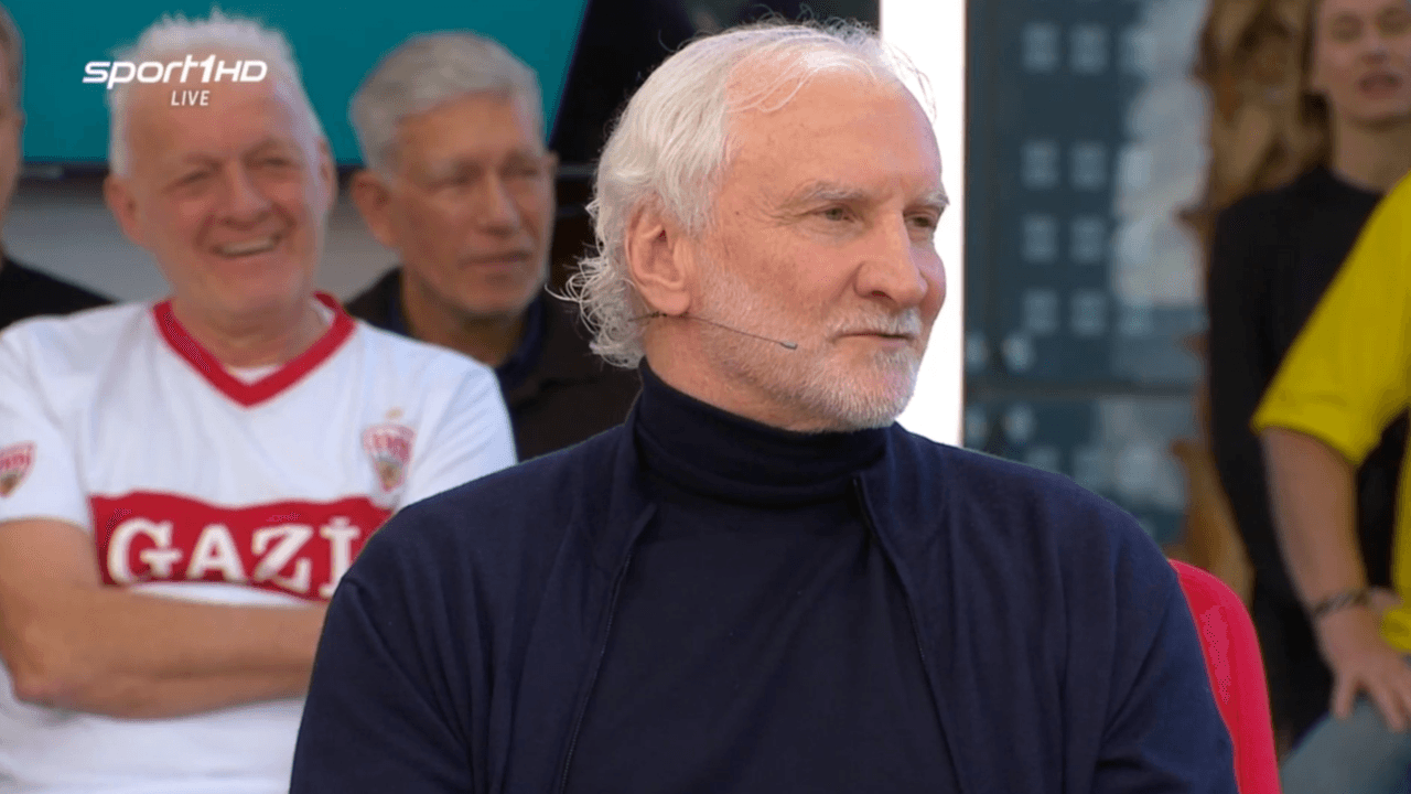 Rudi Völler sitzt mit Rollkragen-Pullover im „Doppelpass“.