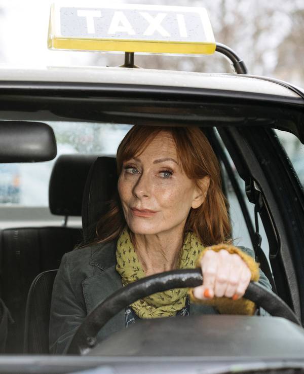 Eine Frau sitzt am Steuer eines Taxis und schaut ungläubig, ein Mann auf dem Beifahrersitz, der eine schwarze Mütze trägt, deutet mit dem Finger.