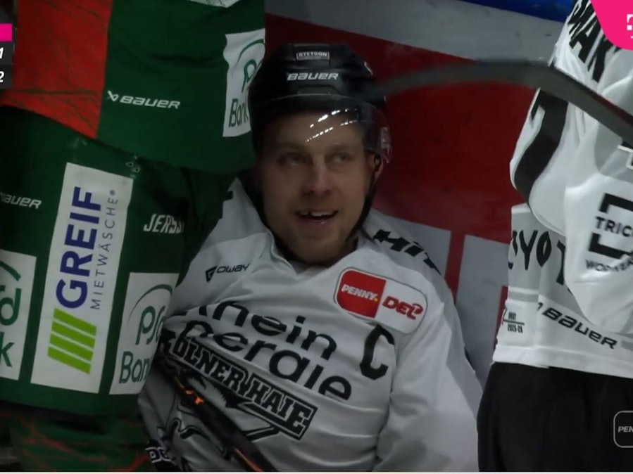 Juhani Tyrväinen sitzt bei einer Rudelbildung an der Bande auf dem Eis und grinst.