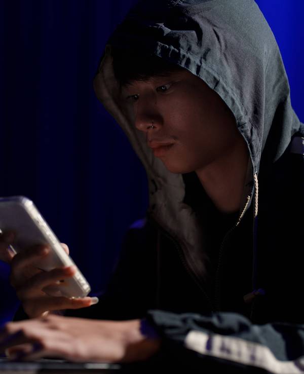 Ein junger Mann sitzt mit Kapuzen-Pullover und Smartphone in der Hand an einem Tisch.