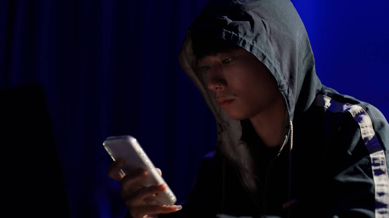 Ein junger Mann sitzt mit Kapuzen-Pullover und Smartphone in der Hand an einem Tisch.