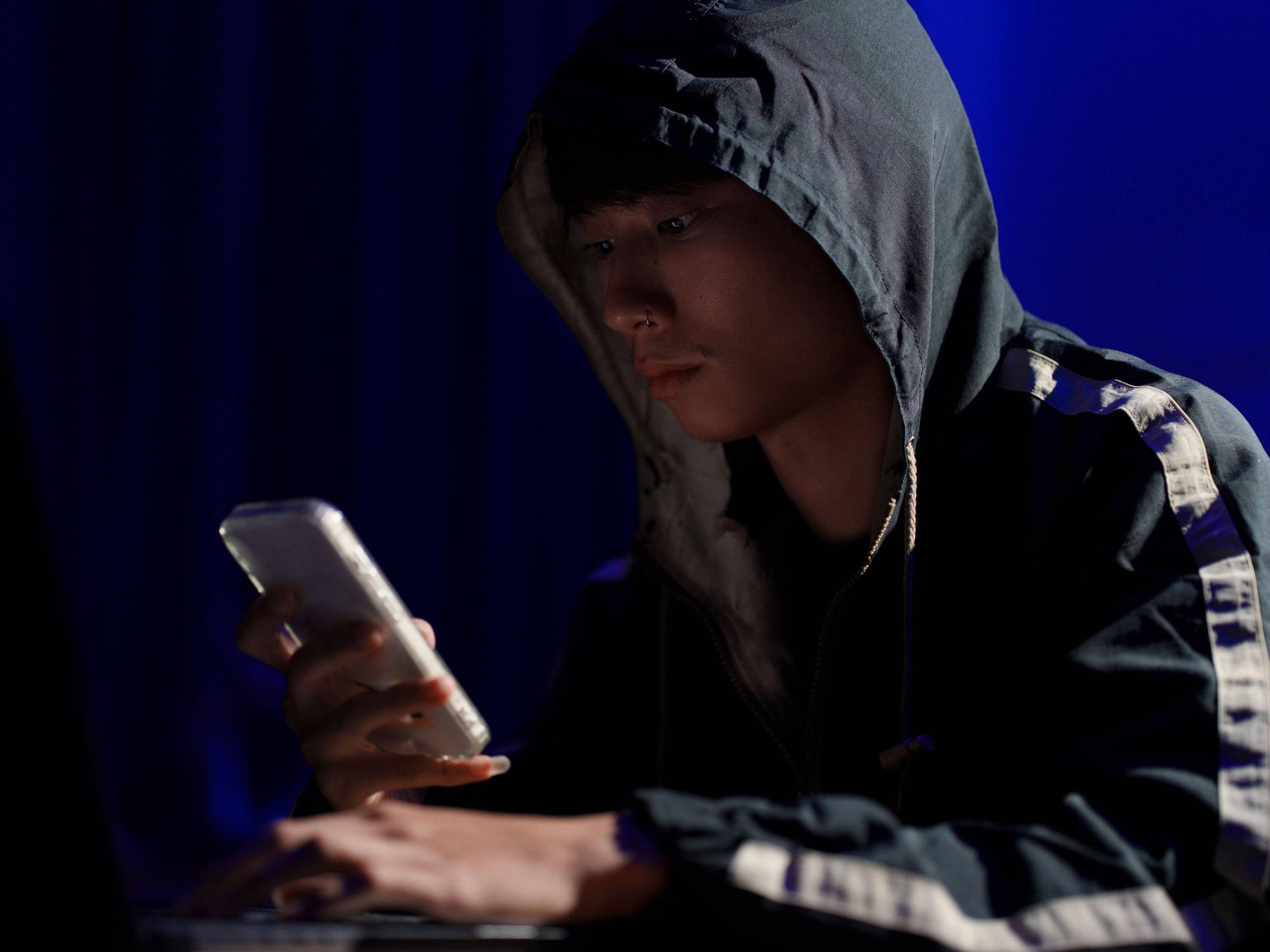 Ein junger Mann sitzt mit Kapuzen-Pullover und Smartphone in der Hand an einem Tisch.