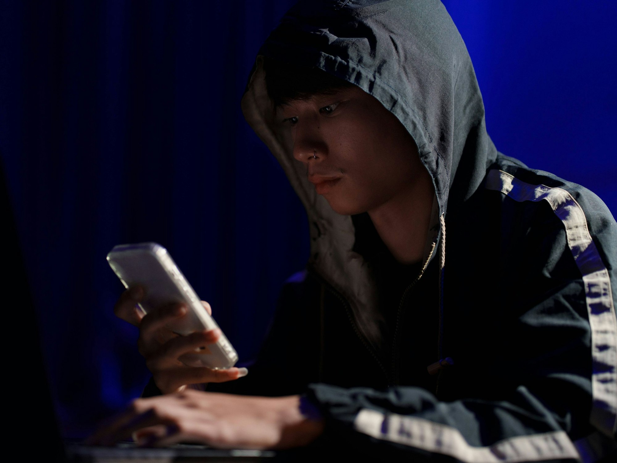 Ein junger Mann sitzt mit Kapuzen-Pullover und Smartphone in der Hand an einem Tisch.