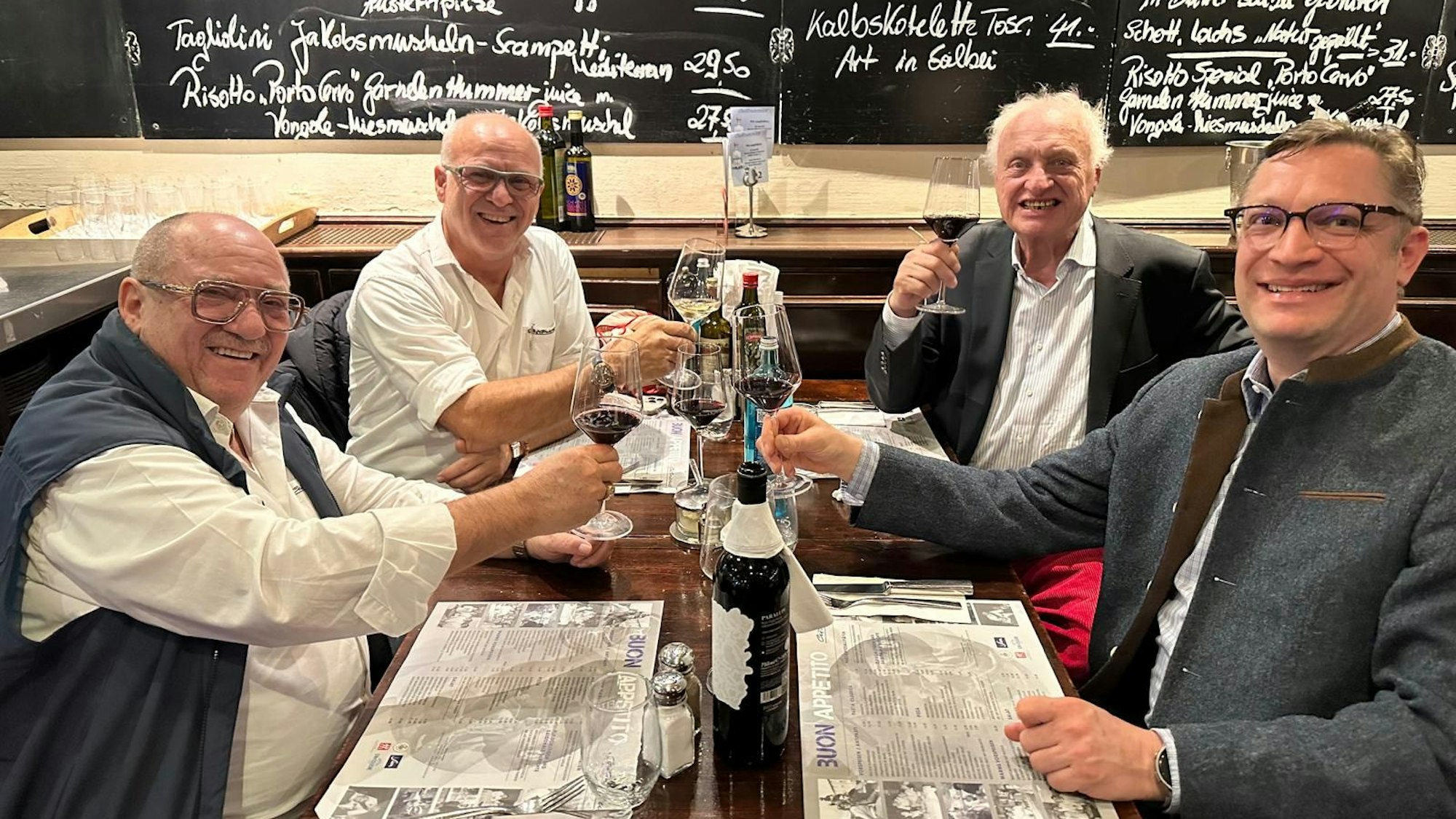 Vier Männer sitzen an einem Tisch und stoßen mit Weingläsern an.