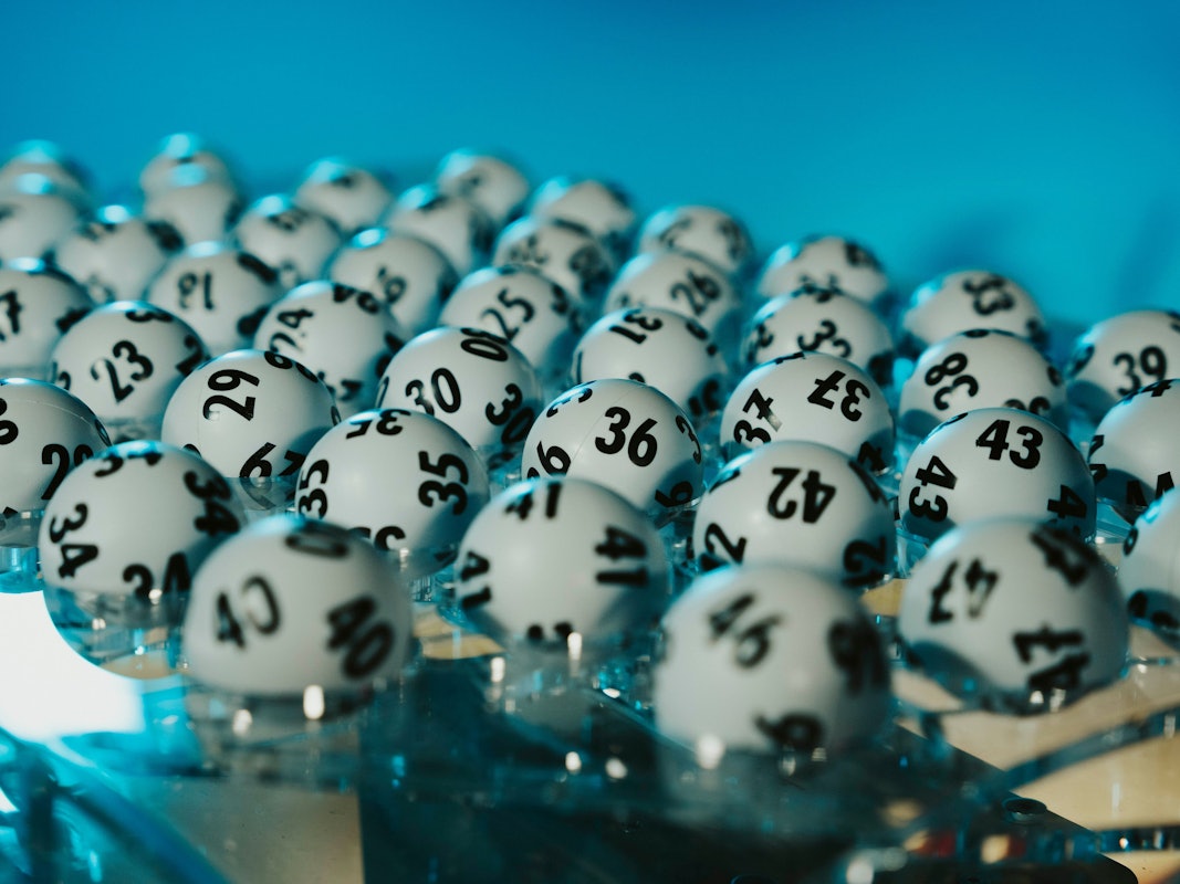 Auf dem Bild liegen zahlreiche weiße, schwarz nummerierte Lottokugeln dicht beieinander in einem transparenten Ziehungsgerät.