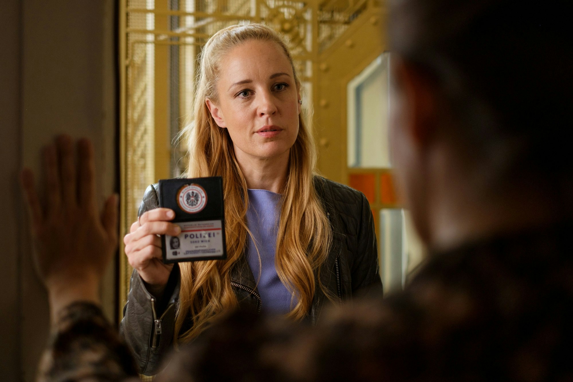 Eine Frau mit langen Haaren steht vor einer geöffneten Wohnungstür und zeigt einen Polizeiausweis vor.