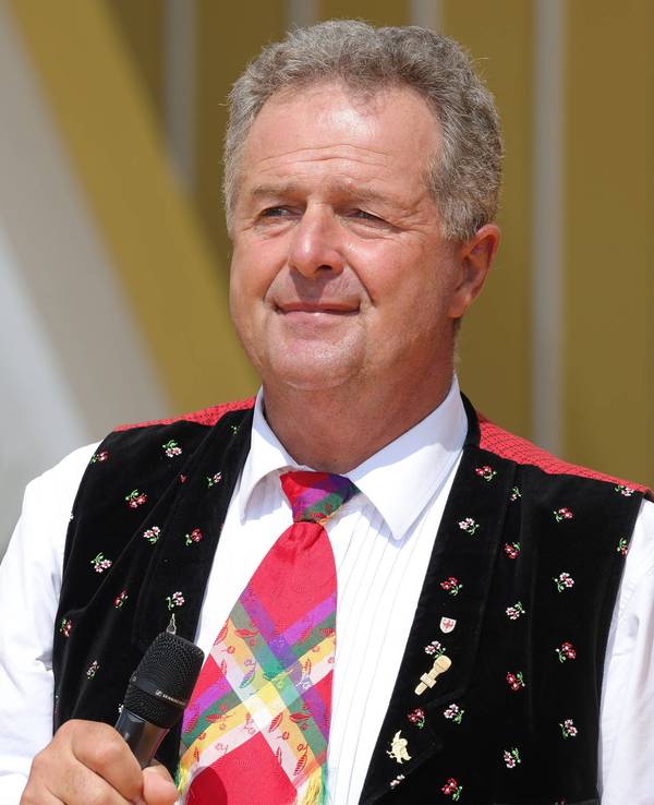 Ein Mann in Trachtenweste mit weißem Hemd und bunter Krawatte hält ein Mikrofon in der Hand und schaut sinnierend in die Kamera.