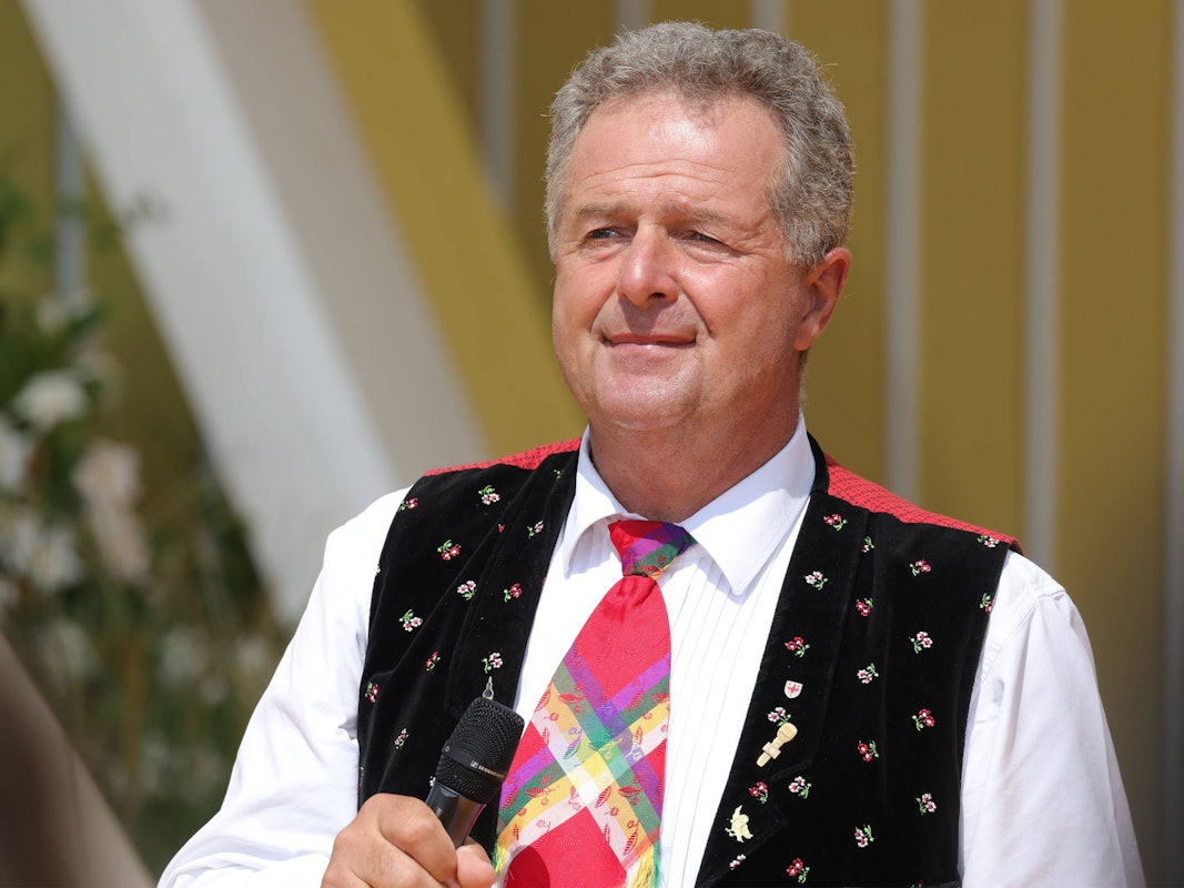 Ein Mann in Trachtenweste mit weißem Hemd und bunter Krawatte hält ein Mikrofon in der Hand und schaut sinnierend in die Kamera.