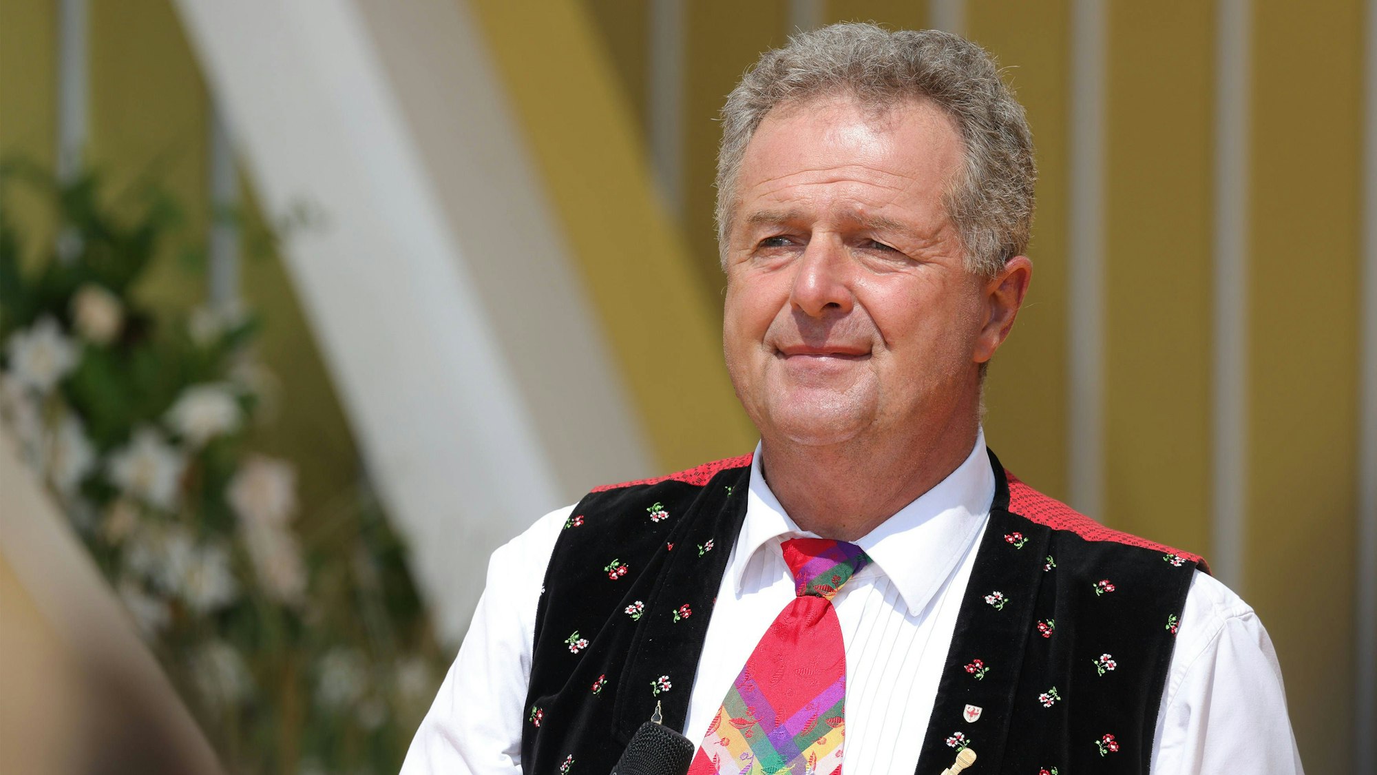Ein Mann in Trachtenweste mit weißem Hemd und bunter Krawatte hält ein Mikrofon in der Hand und schaut sinnierend in die Kamera.