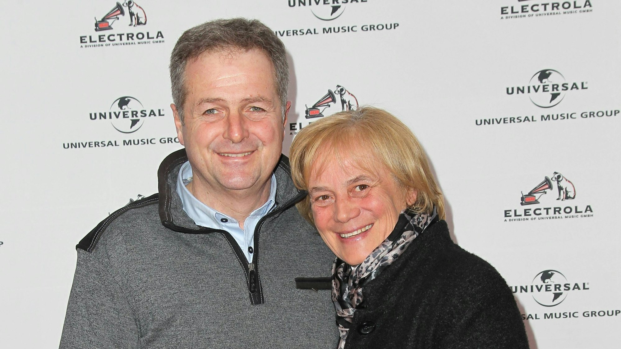 Ein Mann mittleren Alters im grauen Pullover und eine gleichaltrige Frau in einem schwarzen Mantel halten sich im Arm und schauen glücklich.