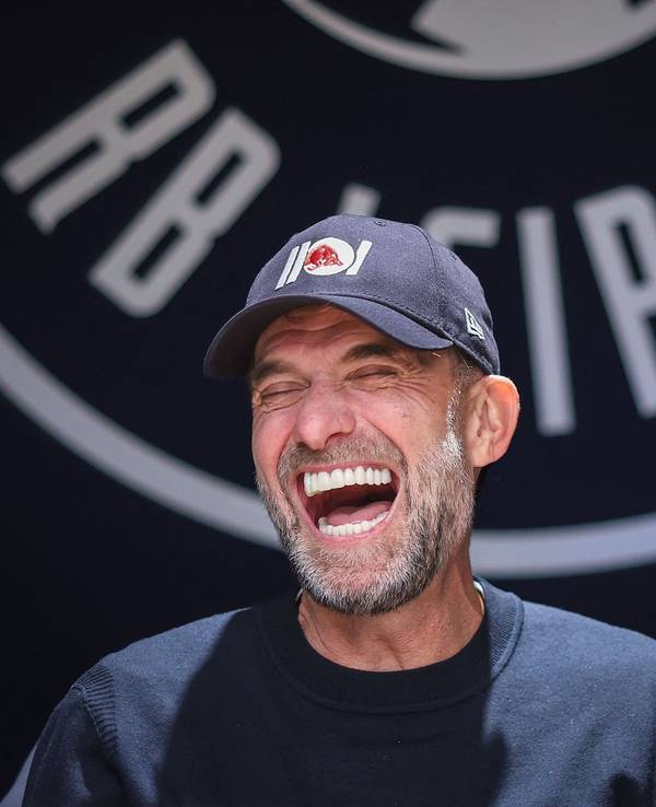 Jürgen Klopp sitzt vor einem großen RB-Leipzig-Wappen, hat eine Kappe auf und lacht.